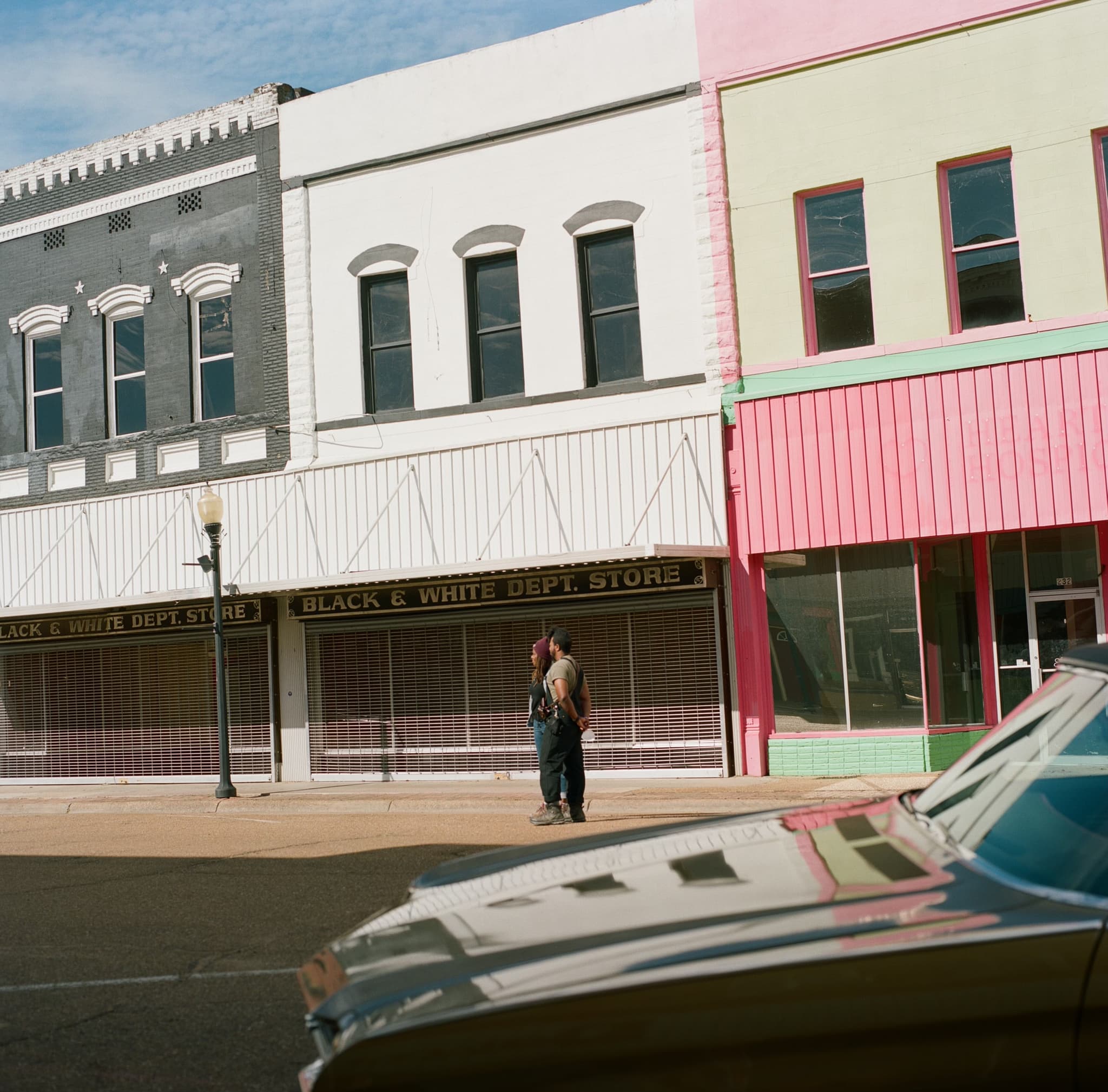 Raven Jackson and Jomo Fray stand in front of colorful storefronts. A sign hanging on one of the store fronts reads “Black and White Dept. Store.”