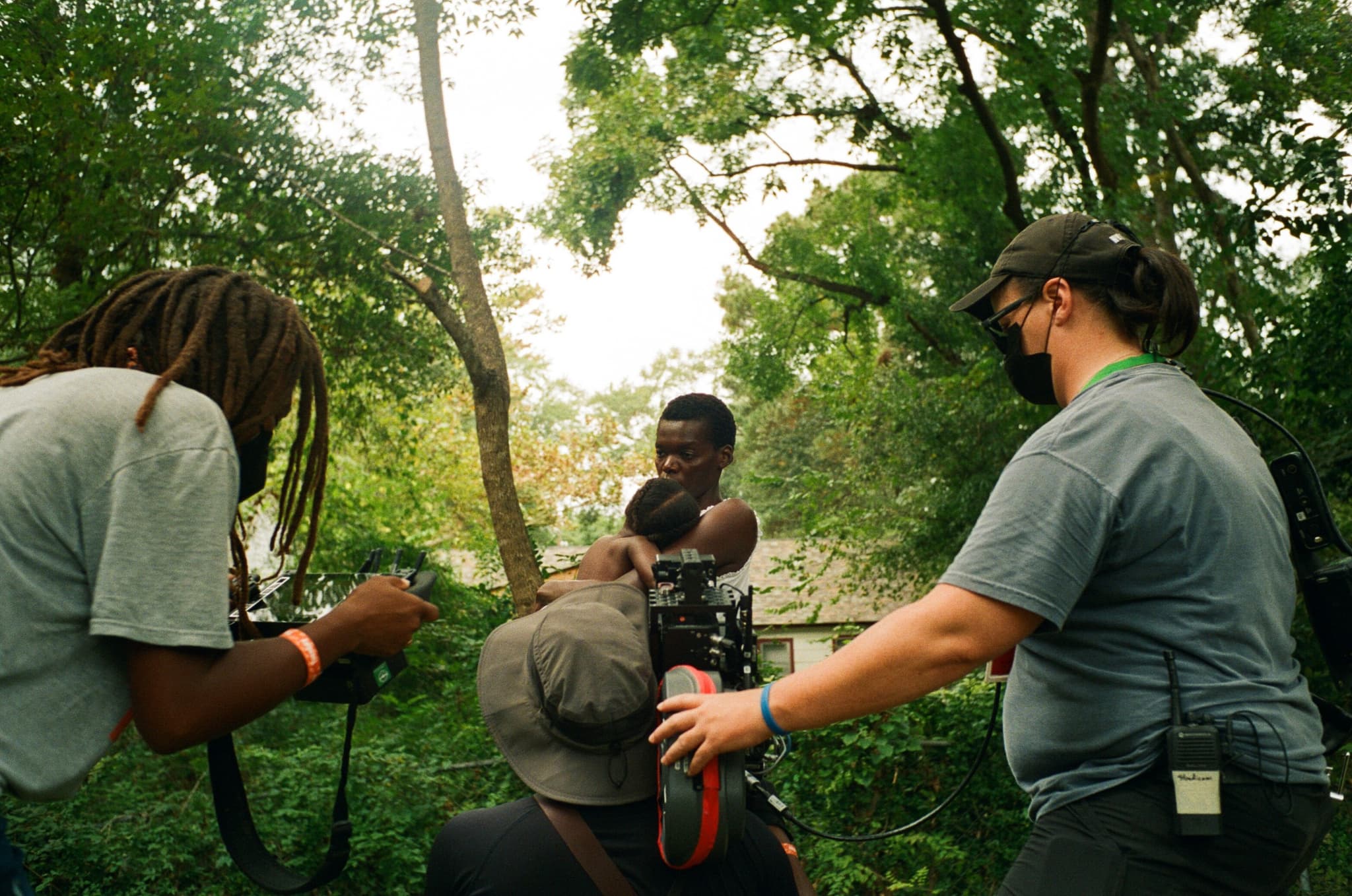 A photograph of Raven Jackson, Jomo Fray, and a camera assistant (from left to right) on the set of All Dirt Roads Taste of Salt along with two actors from the film. They are in a lush green forested area. Jackson holds a monitor and faces away from the camera. Fray holds the camera on his shoulder and faces away from the camera. The camera assistant reaches for the back of the camera to support.