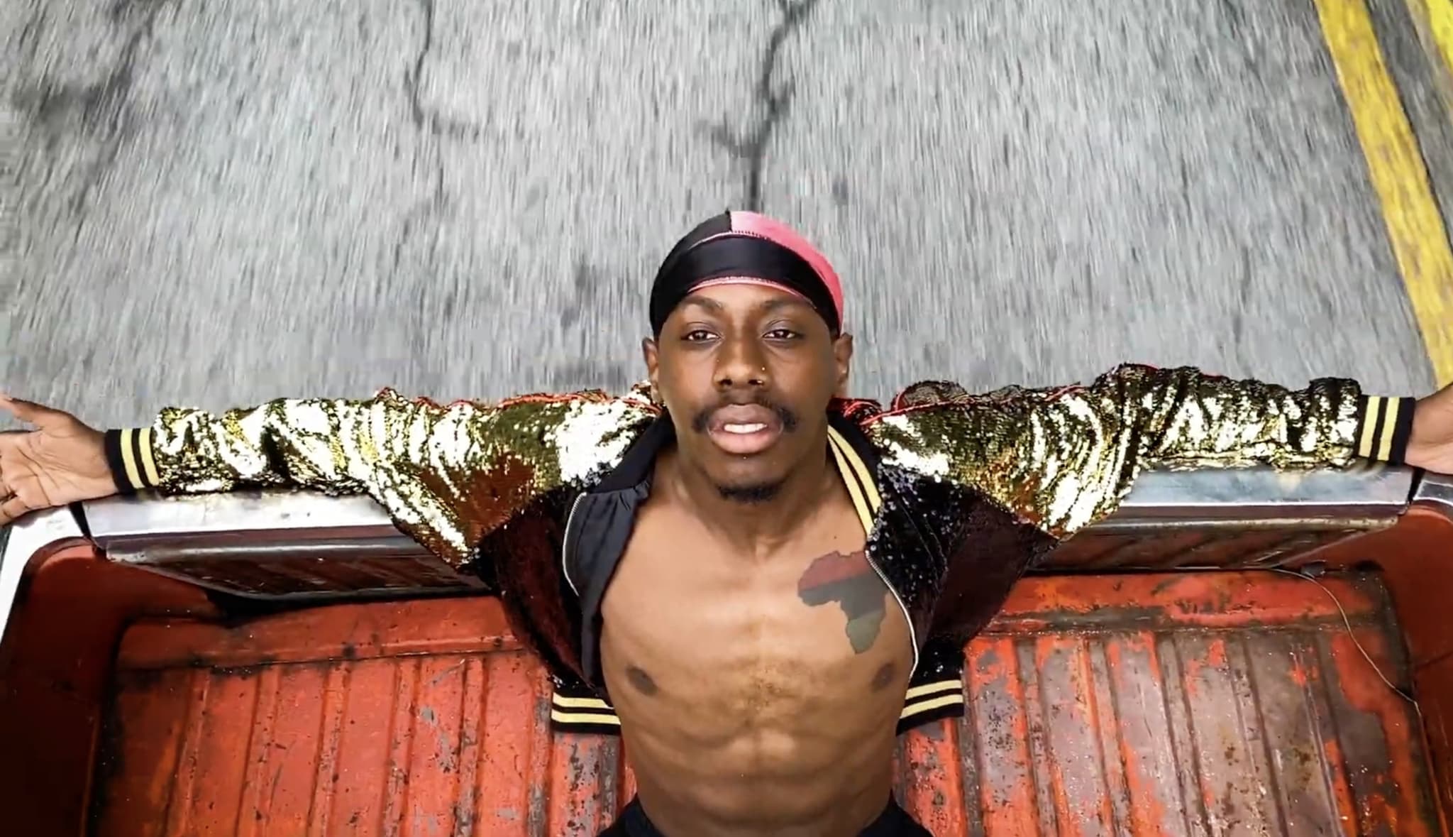 A photo of a Black man wearing an unzipped gold and black varsity jacket sitting back in the back of a red pick-up truck. His arms are outstretched and he is leaning his head back and looking towards the sky.