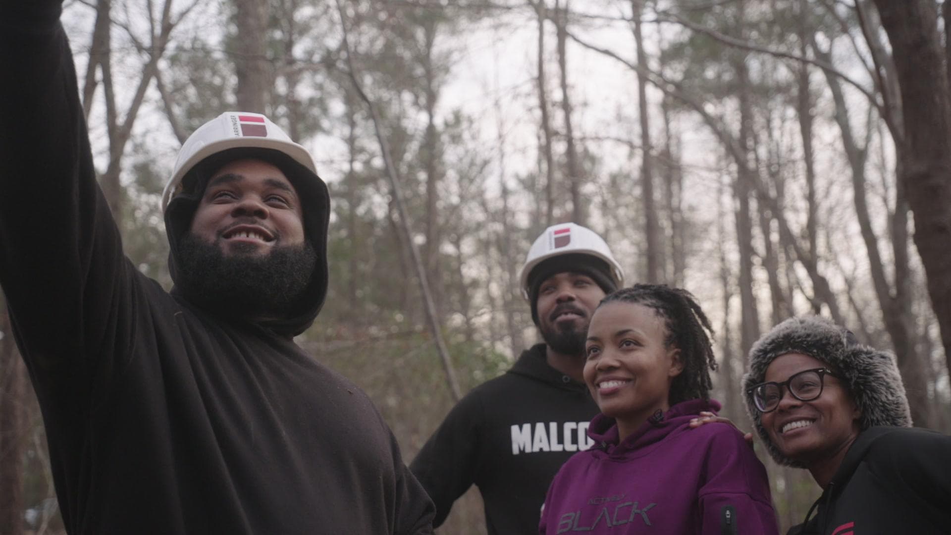 A still from Family Tree shows four Black people standing in a forest. Two are wearing hard hats. They are looking up at something and smiling.