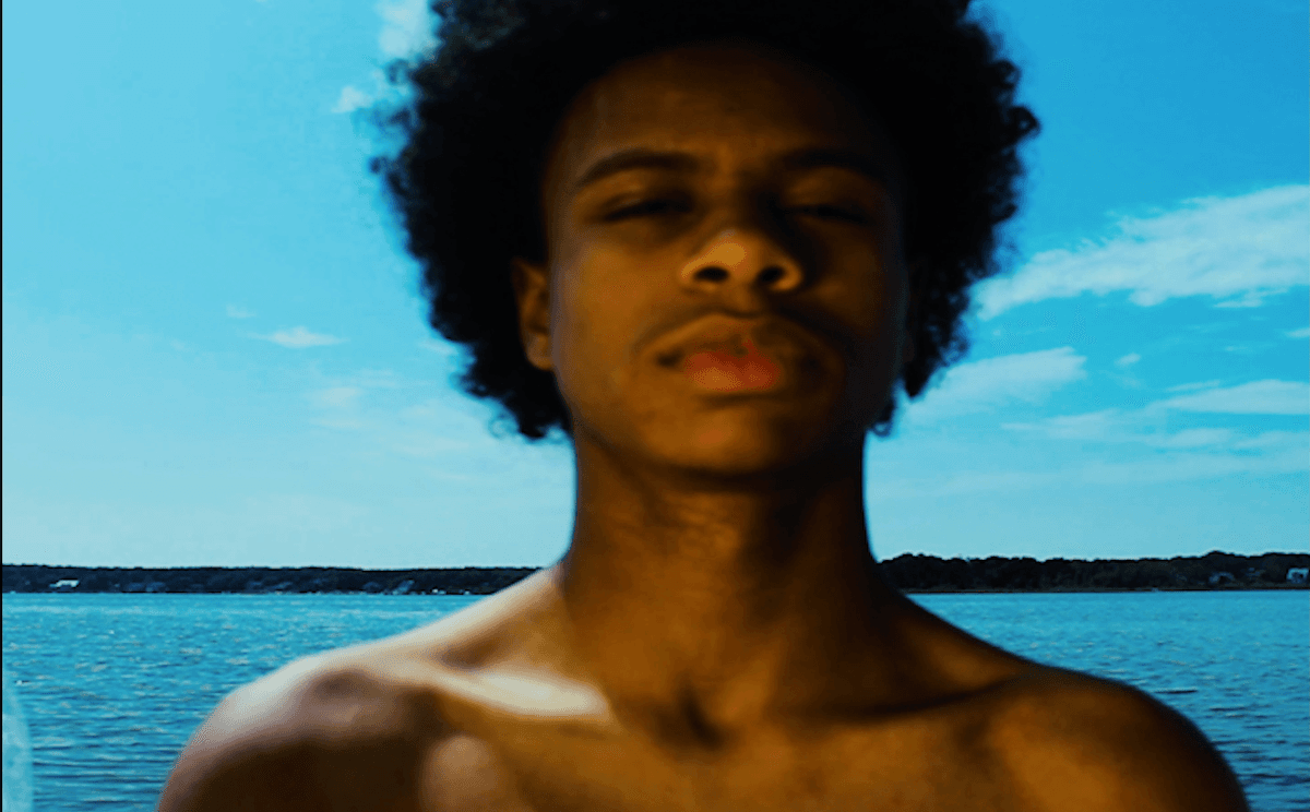 A still shows a man with an afro standing in front of a body of water. His eyes are squinting.