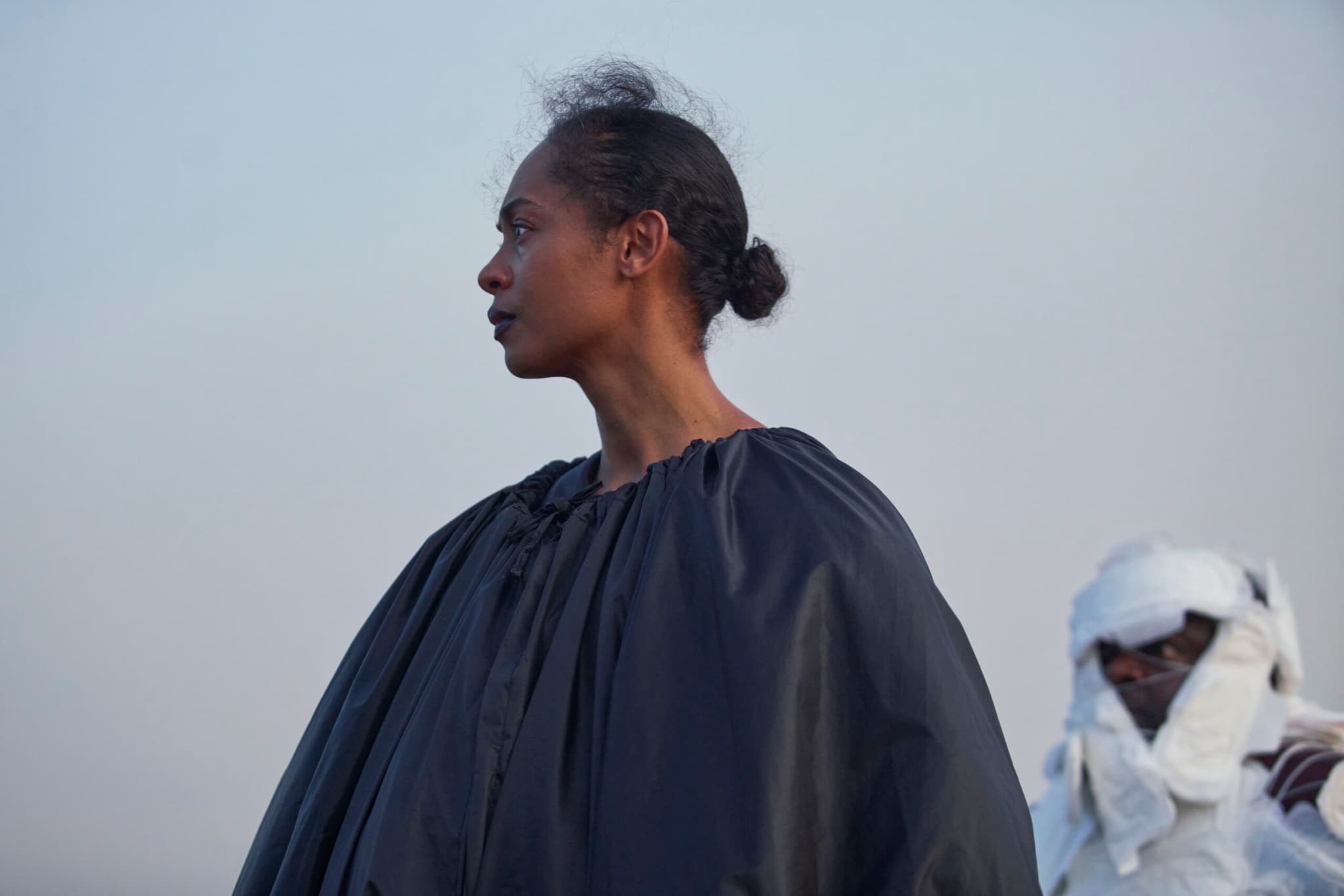 A still from on Becoming A Guinea Fowl shows the profile of a woman. She is wearing a large black cape. She is looking off into the distance, her expression is concerned.