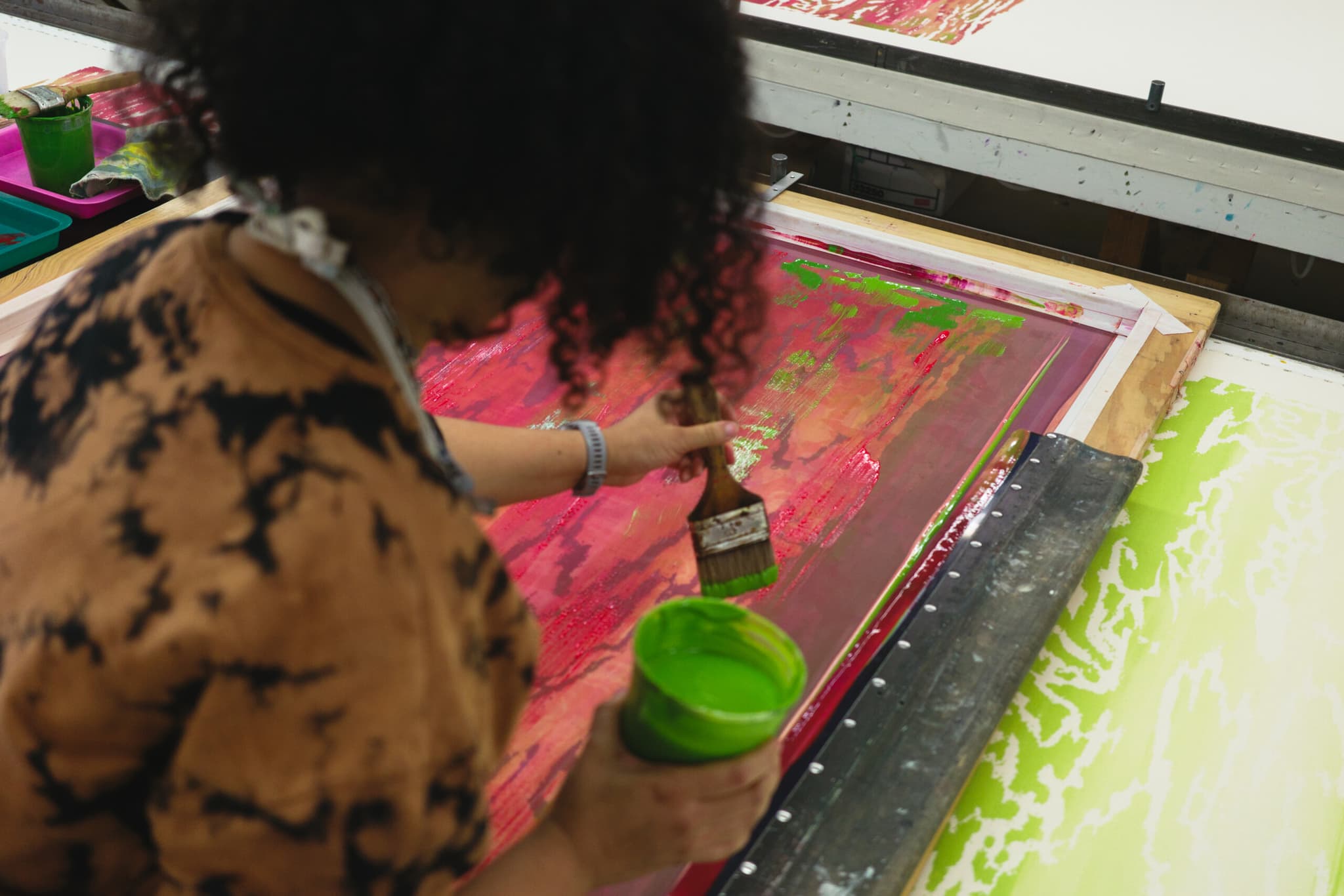 A photo taken over the shoulder of an artists with brown curly hair painting a canvas while holding a cup of bright green paint.