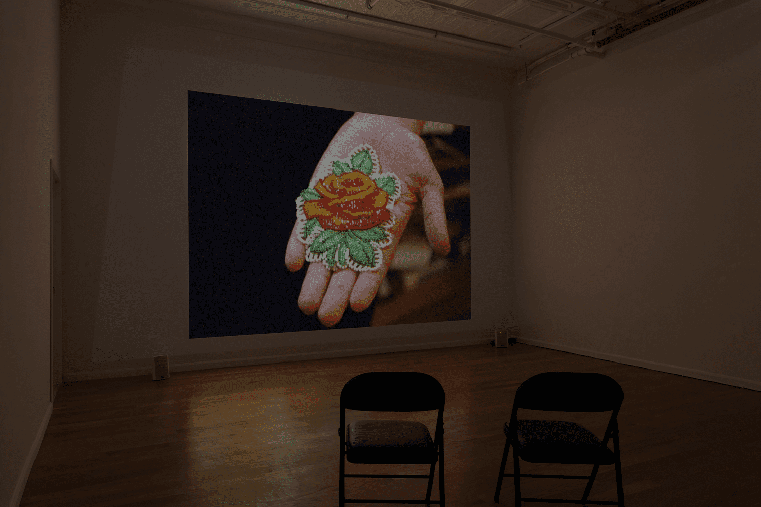 A photo of an empty gallery space where a film is showing. In the film an outstretched hand is showing a sewn object, which appears to be a flower.