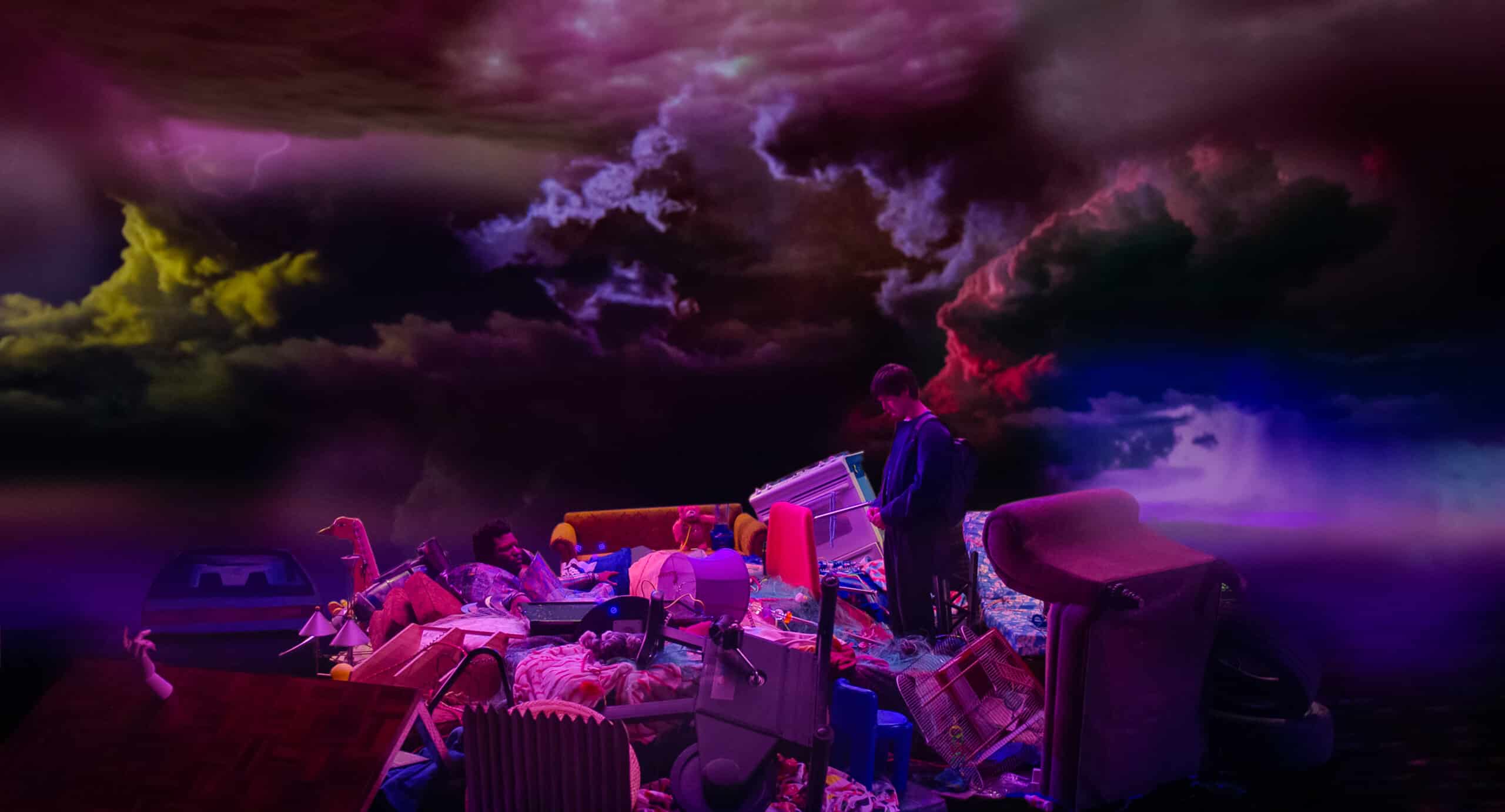 A still image from “Problemista” (2023) featuring a young man stares at a pile of his belongings. Above him is a sky with stormy clouds.