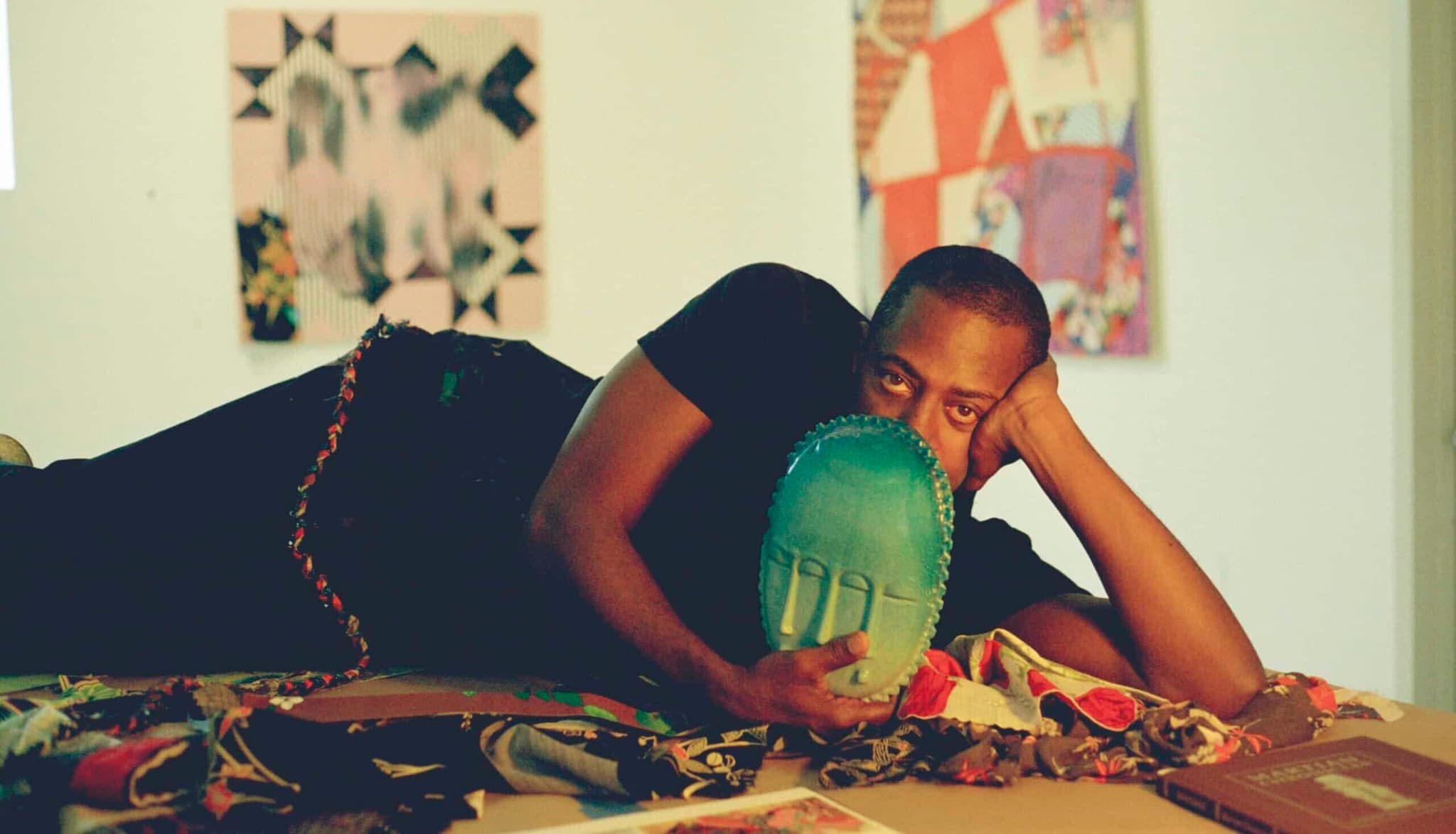 Sanford Biggers lying down on his side with one hand holding his head up, and the other holding an art piece in the shape of a sculpted teal mask that covers the bottom half of the face.