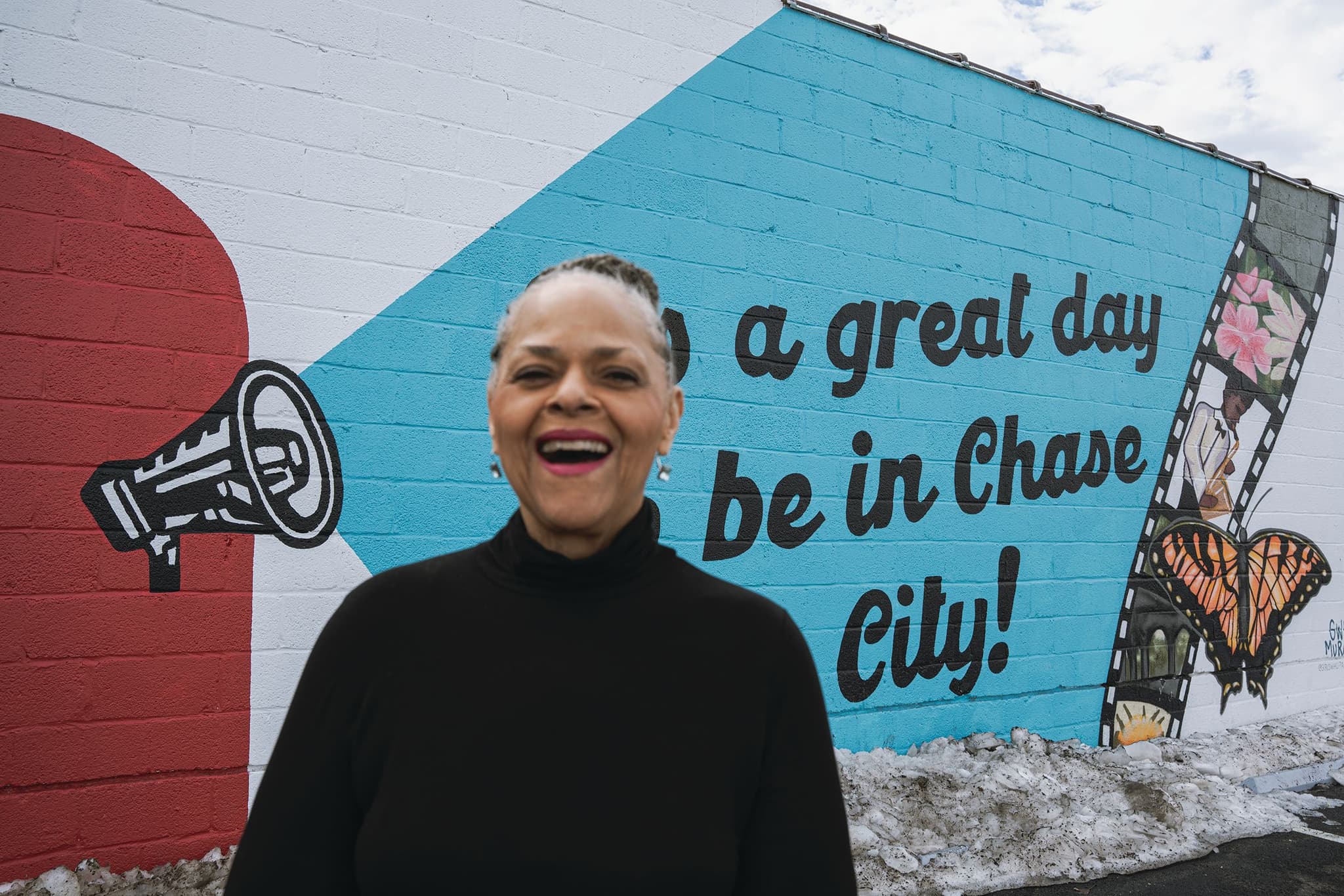 Beverly Wood is laughing in front of a “It's a great day to be in Chase City” wall art.