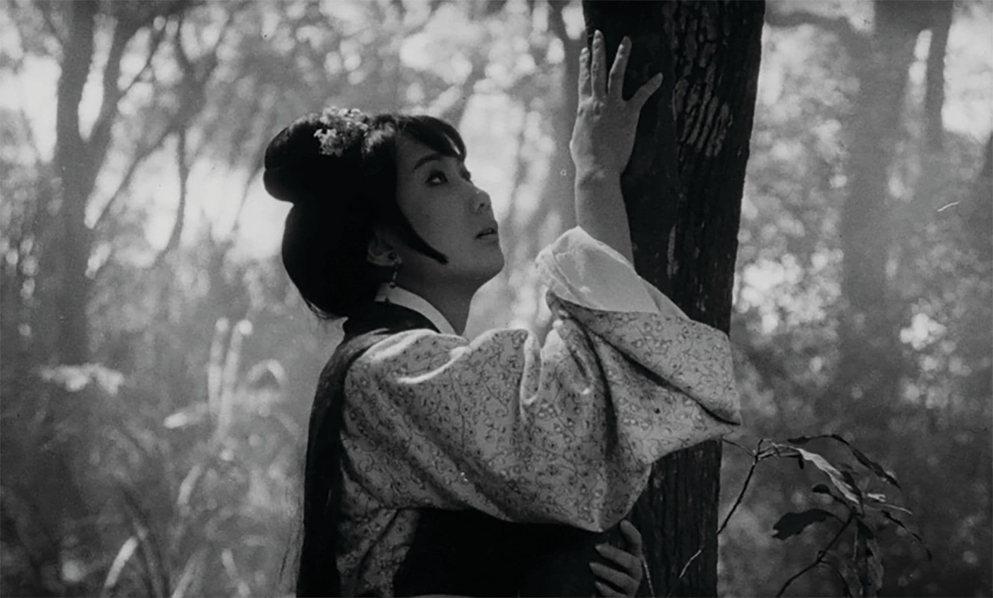 A black and white still from the film, The Arch, where a woman is hugging a tree and looking up.
