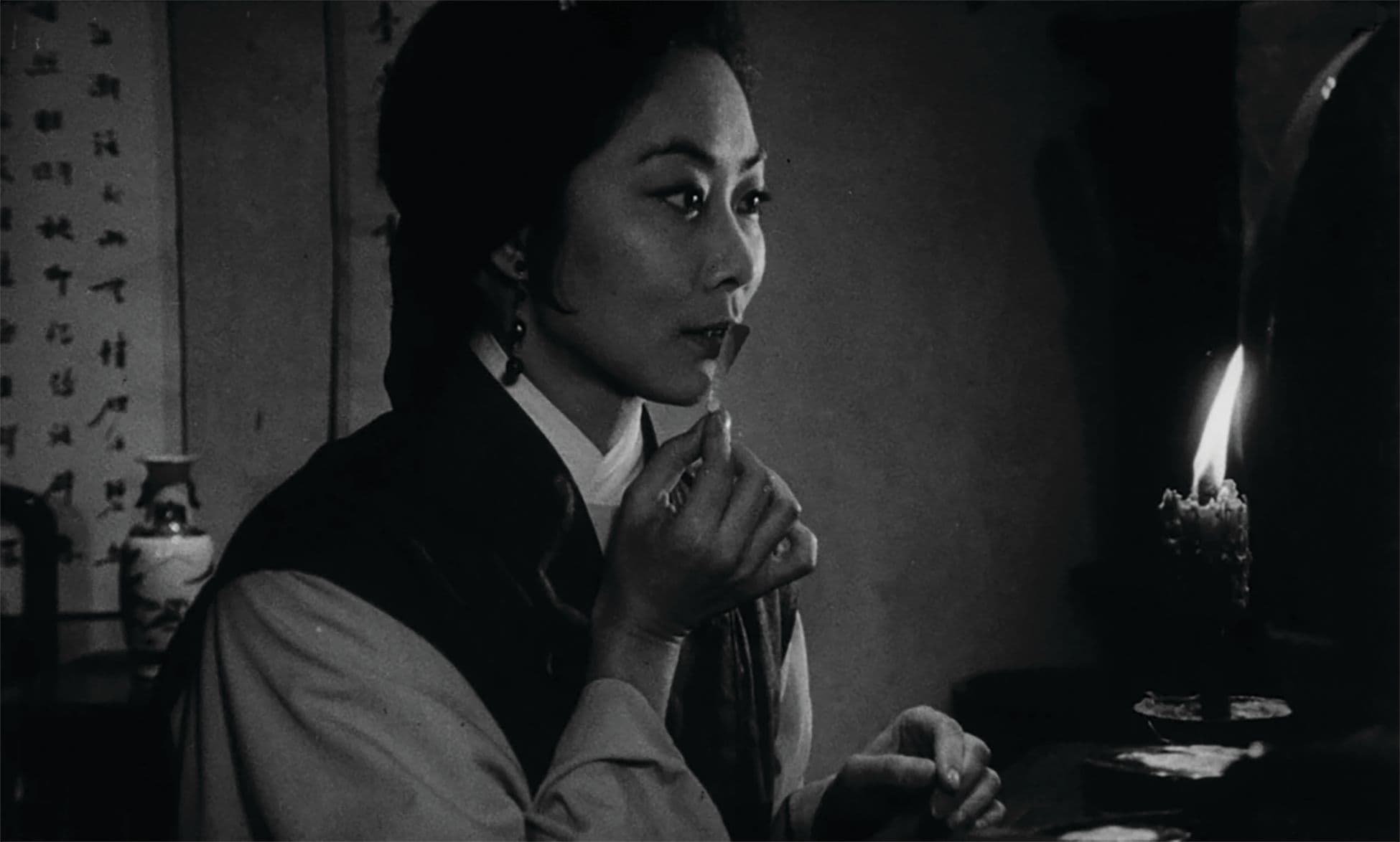 A black and white still of a woman applying her lipstick in front of a mirror.