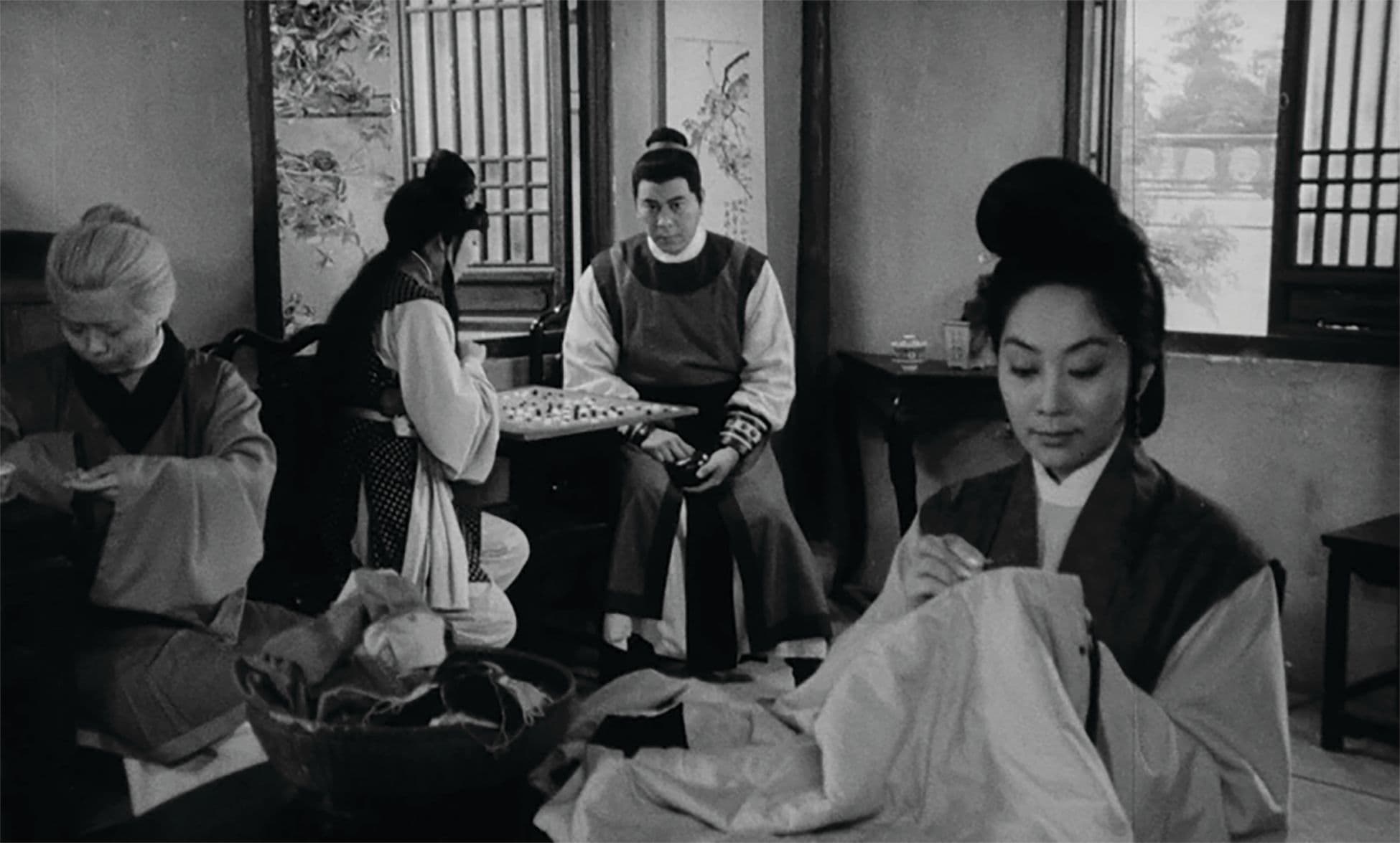 A black-and-white still of people gathered in a living room: two people in front are sewing, while others in the background are playing board games.