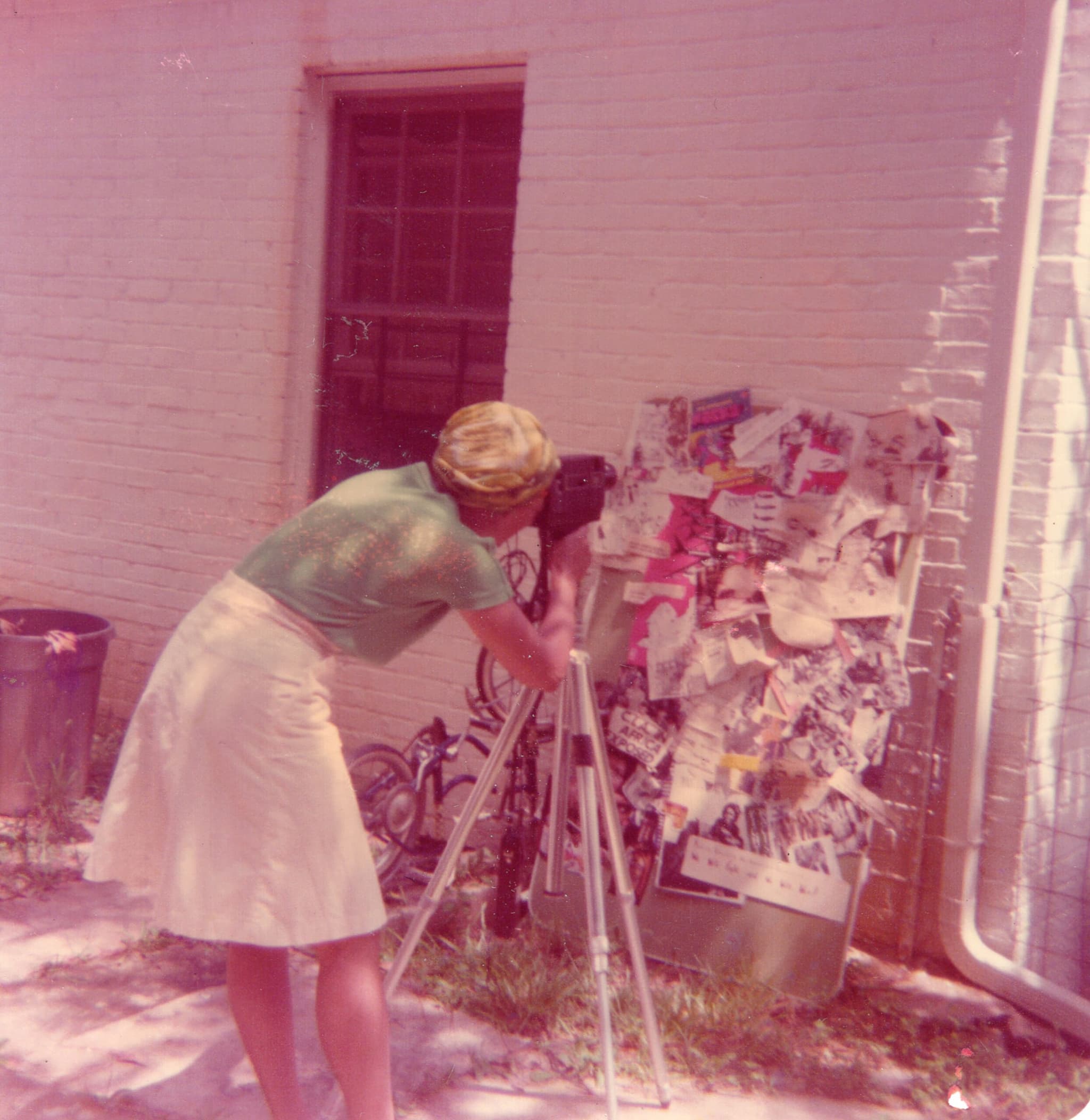 Toni Cade Bambara is taking a picture of a photo collage that’s leaning on the outside of a building. Her back is facing towards the camera, and she is using a tripod.