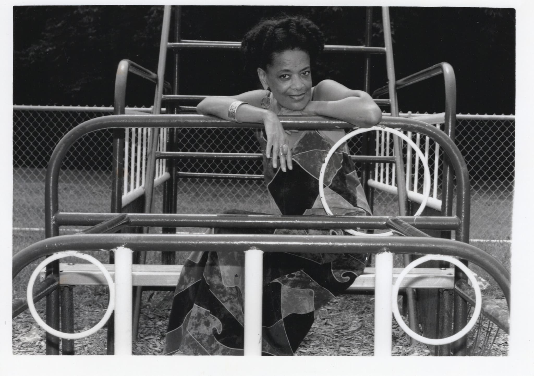 Toni Cade Bambara is sitting on a bench with her hands on the rail, and her face turned slightly to the right.