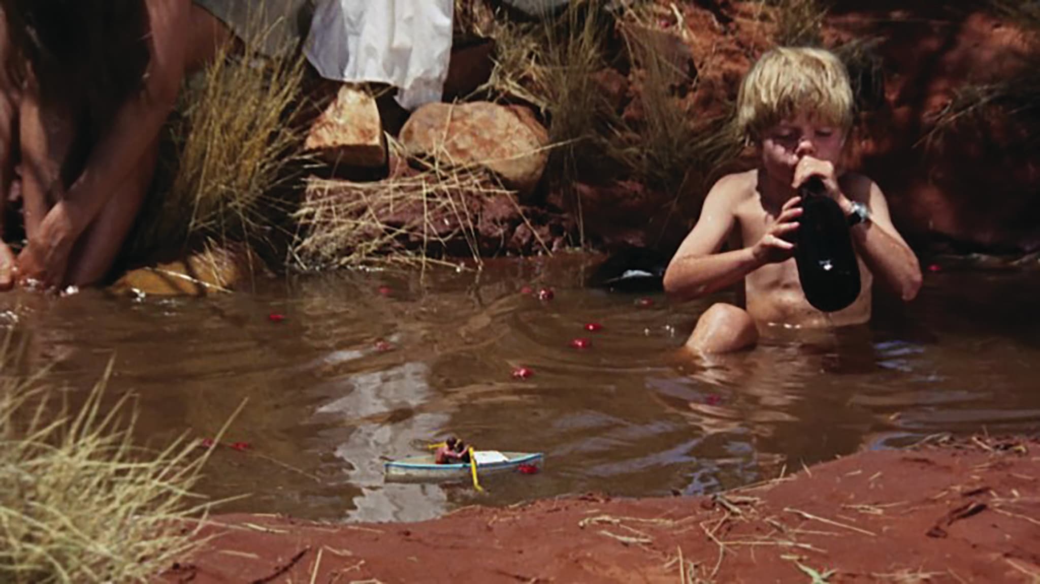A still from Walkabout, where a little boy is sitting in murky water with a water bottle in hand. To his left is someone washing their feet in the same water. A white toy boat floats next to him.