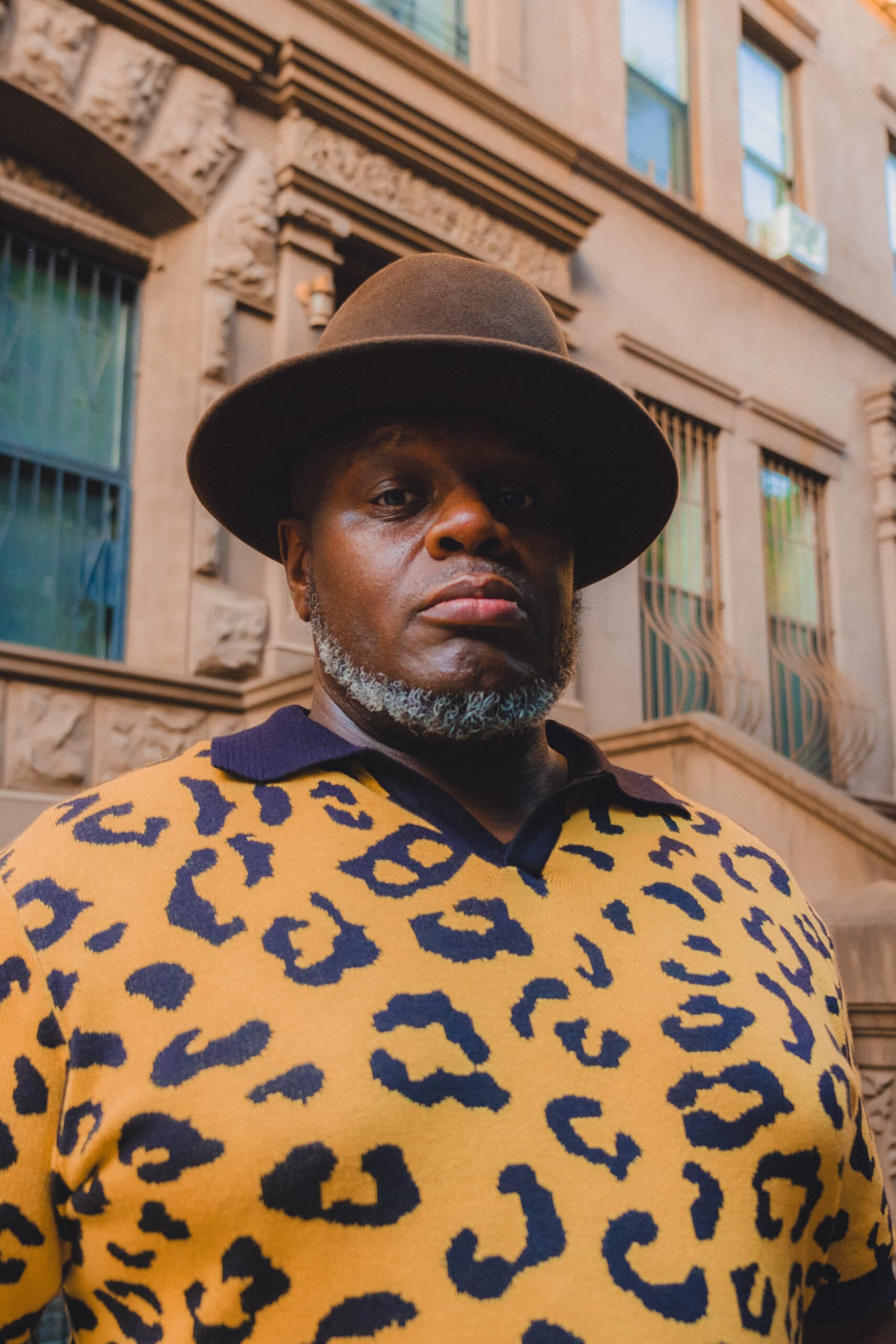 A headshot of Anthony Tidd. He is a Black man, he is wearing a brown-brimmed hat and a leopard print polo shirt.
