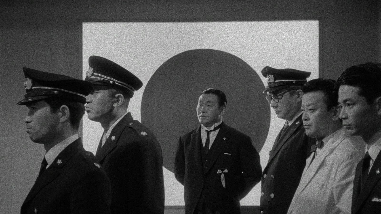Black-and-white film still of a stern-looking man in a dark formal suit with a white collar and a ribbon pinned to his chest, standing in front of a large dark circle on a pale wall. Two uniformed officers stand on either side of him.