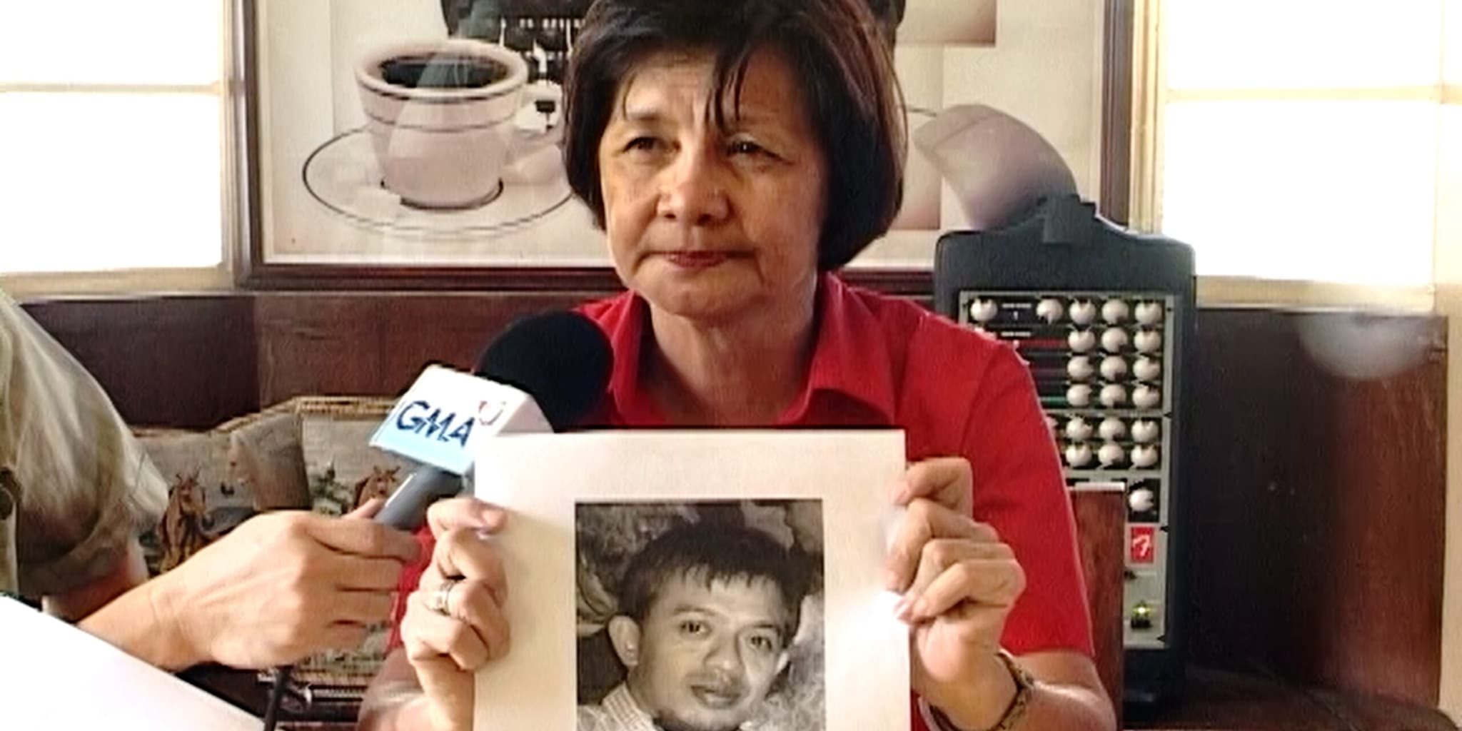 A woman in a red shirt holds up a black-and-white photo of a young man as a reporter extends a GMA microphone toward her.