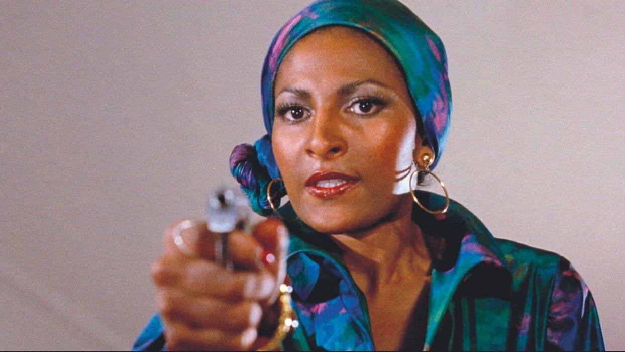 Film still of a Black woman in a blue head wrap and gold hoop earrings aims a revolver at the camera.