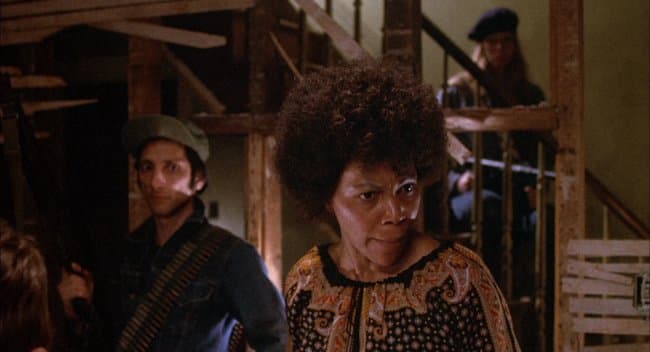 Film still of a Black woman with a full afro and a patterned top gazing intently to one side, a man in a knit cap beside her in the shadows.