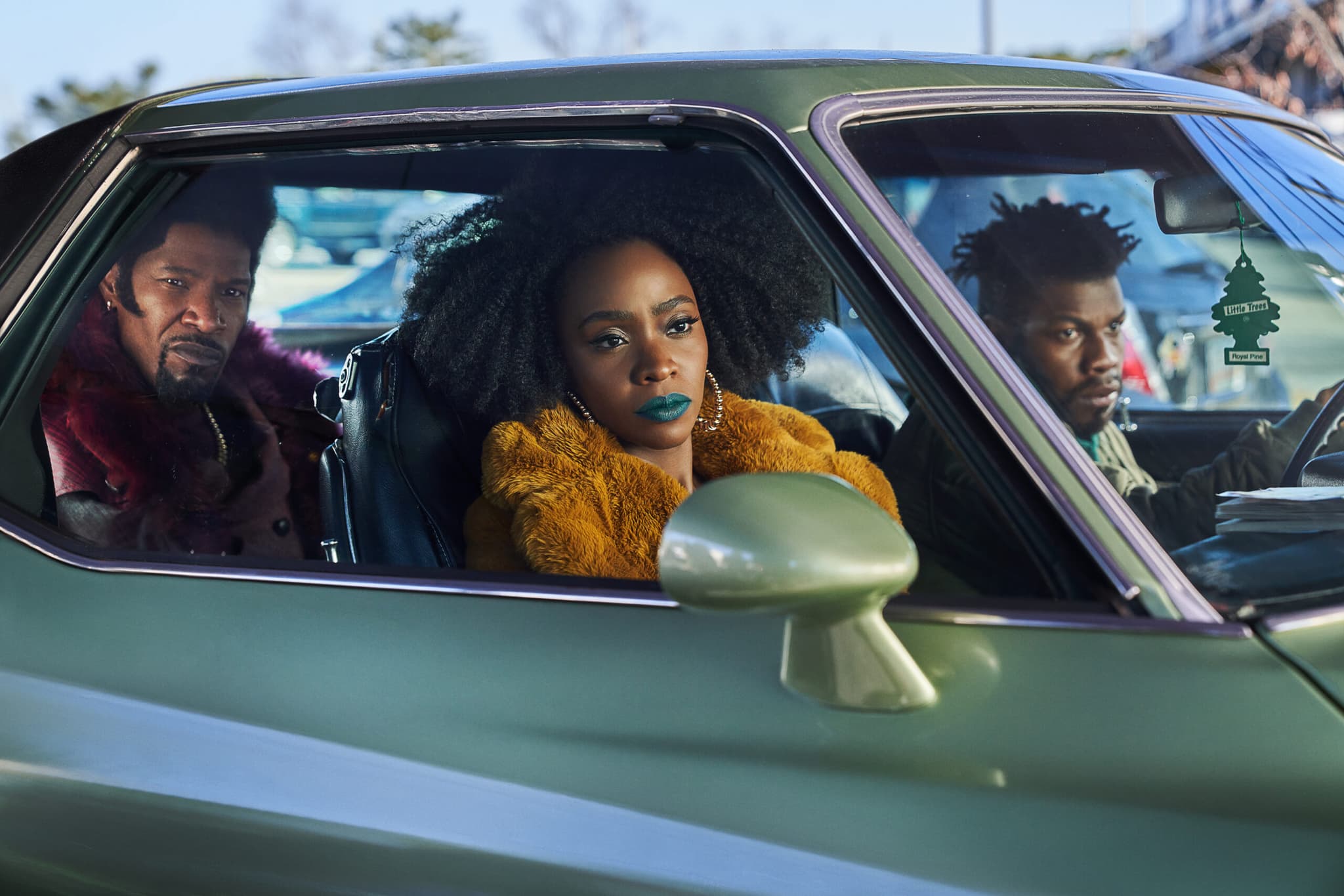 A young Black woman and man are in the front seat of a car. She wears blue lipstick and stares out the window. A young Black man is in the back of the car.