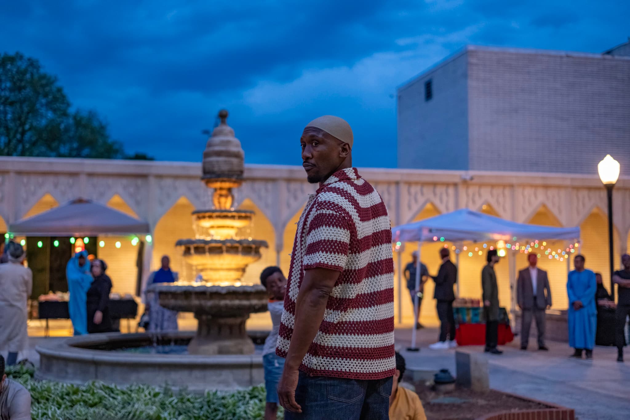 A Black man stands in the foreground and stares at something to the side and behind him. In the background, a young man stands near a lit fountain and stares in the same direction. There are people standing near the fountain.