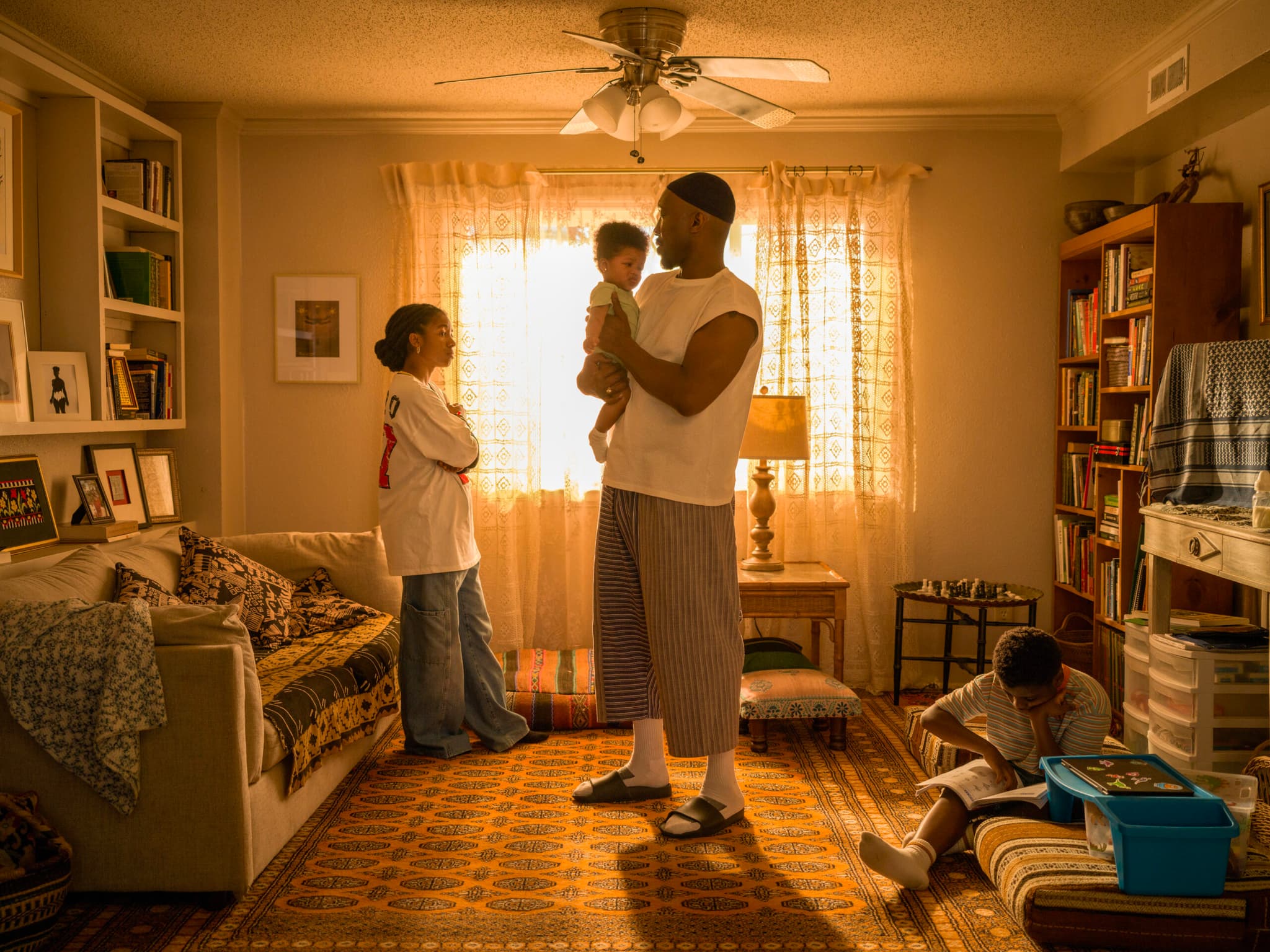 In a sun-lit living room, a Black man holds a young child and looks at a young woman across from him, who stands with her arms crossed. A young Black man sits next to the man, reading.