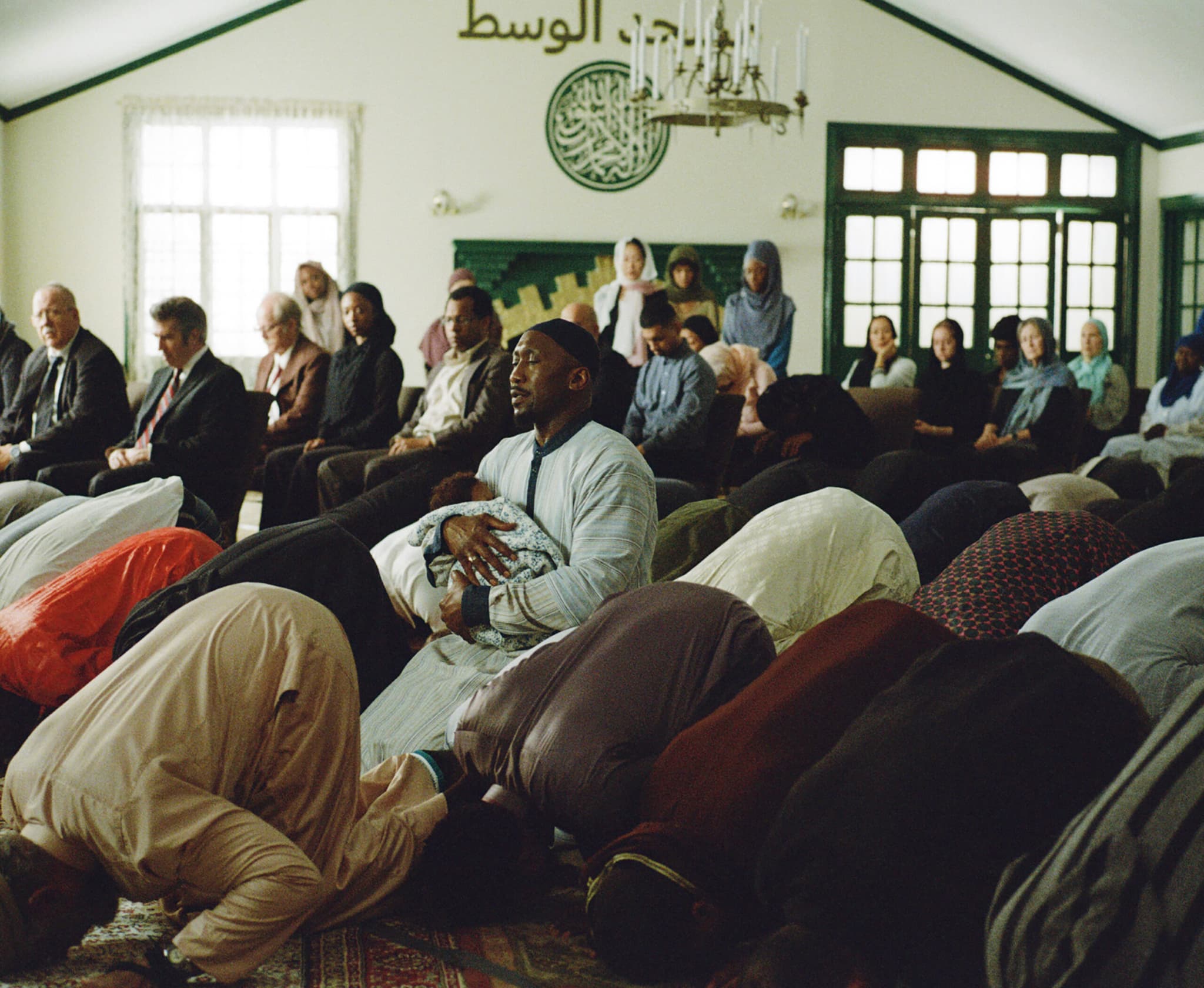 In prayer and eyes closed, a Muslim man kneels upright with a baby in his arms, while others are prostrate in prayer around him.