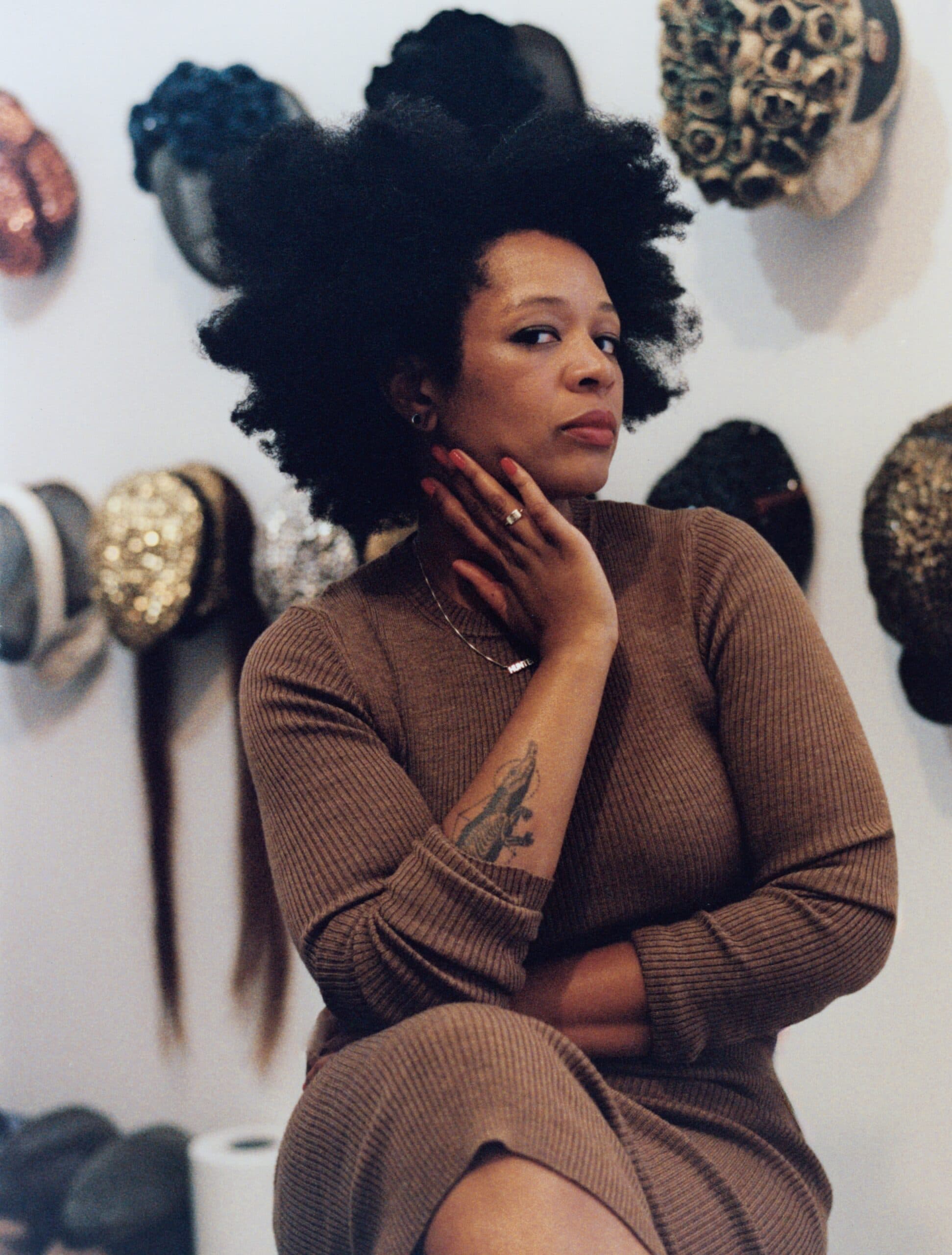 Portrait of Allison Janae Hamilton, a Black woman with full, dark natural hair, wearing a brown turtleneck and a thin chain necklace. She rests one hand against her jaw, showing a ring and a forearm tattoo, and looks at the camera with a steady expression.