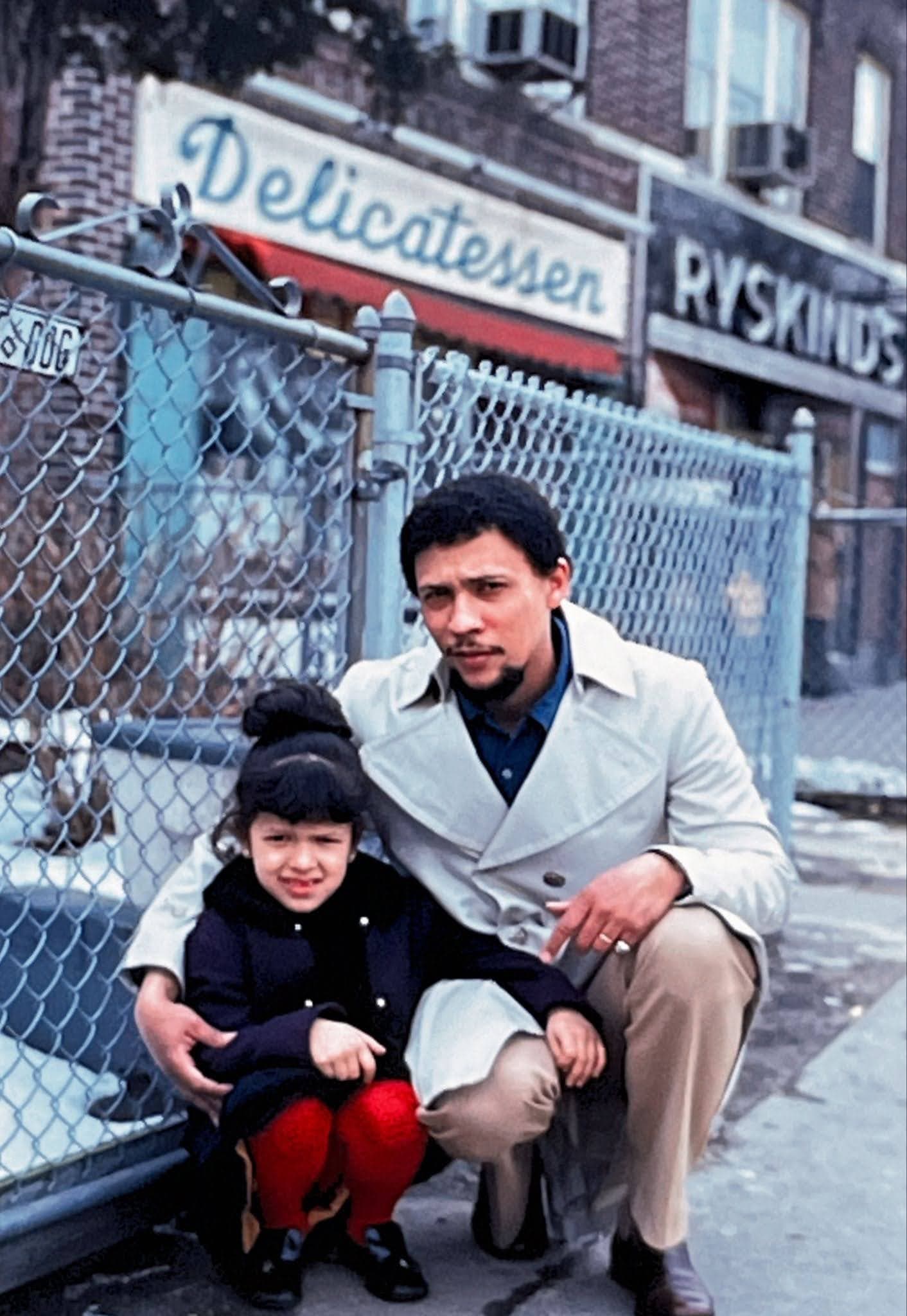 A Black man in a white coat crouches down beside a young girl in a black coat, his arm around her.