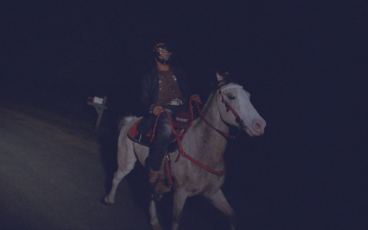 A still from When I Get Home. A Black man is riding a white horse down a road at night.
