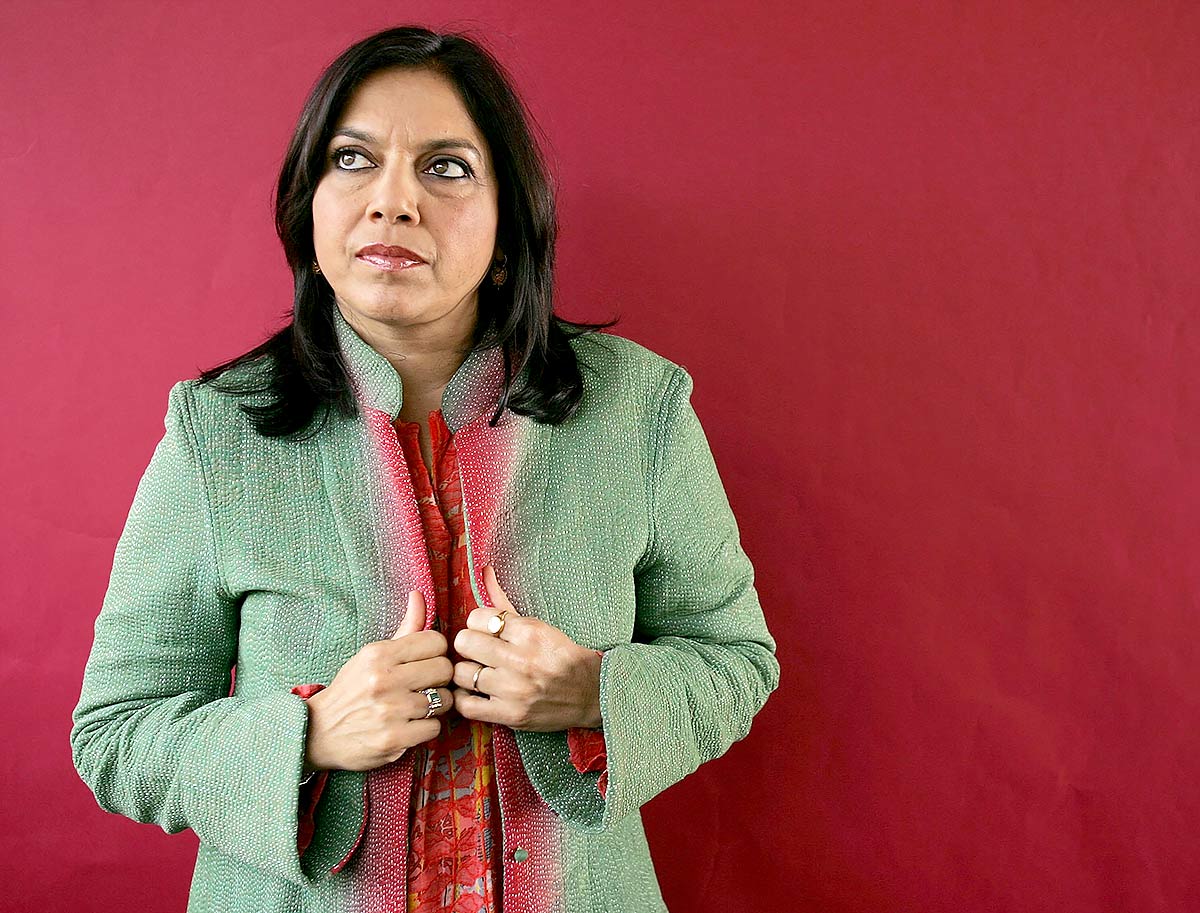 A headshot of Mira Nair, she is standing in front of a red wall. She is wearing a green and red coat and she is looking off to the side with a contemplative expression.