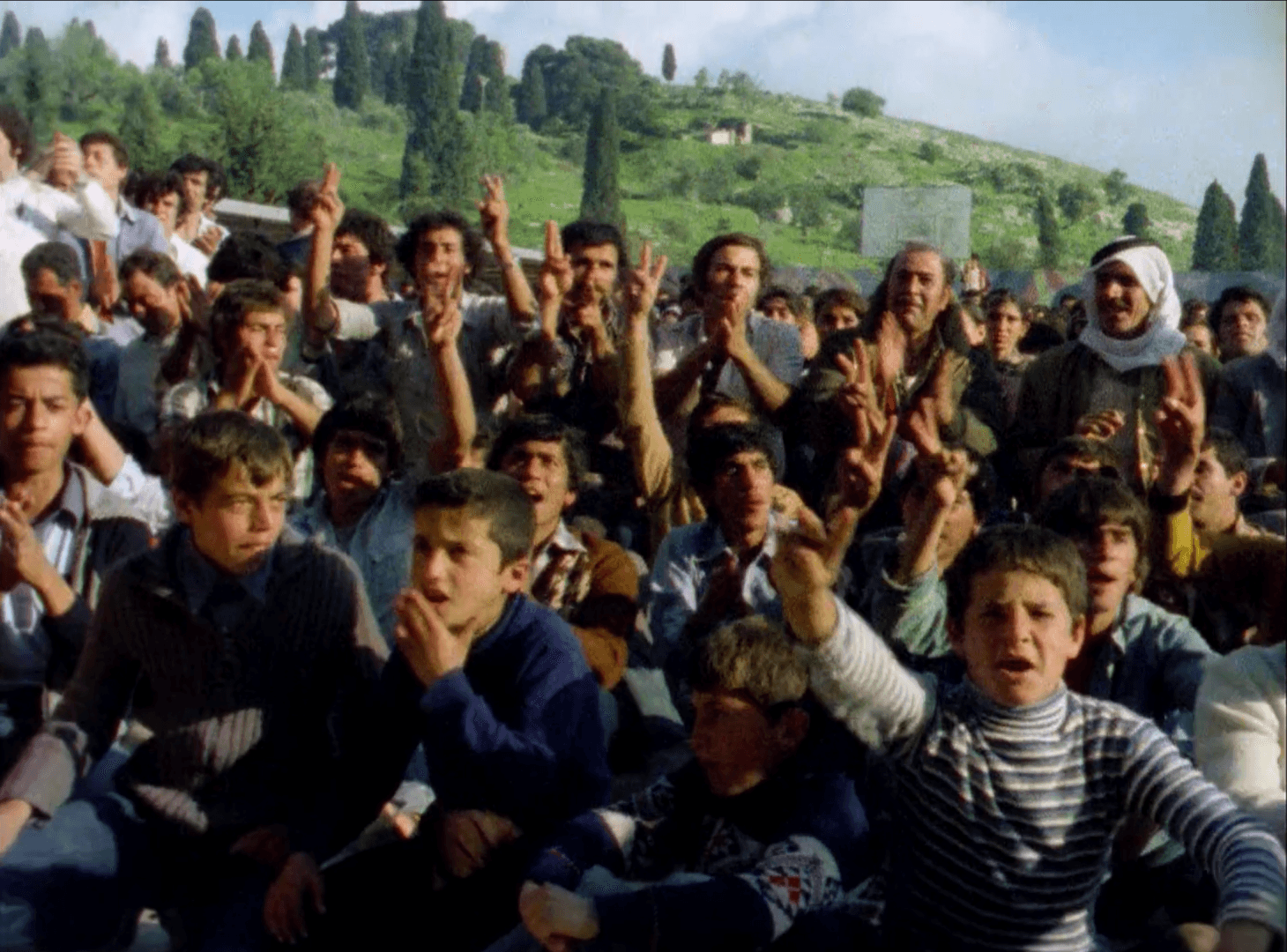 Still from Yom al-Ard (2019), dir. Monica Mauer, showing a crowd of children in Palestine raising their hands and cheering. Behind them are green hills.