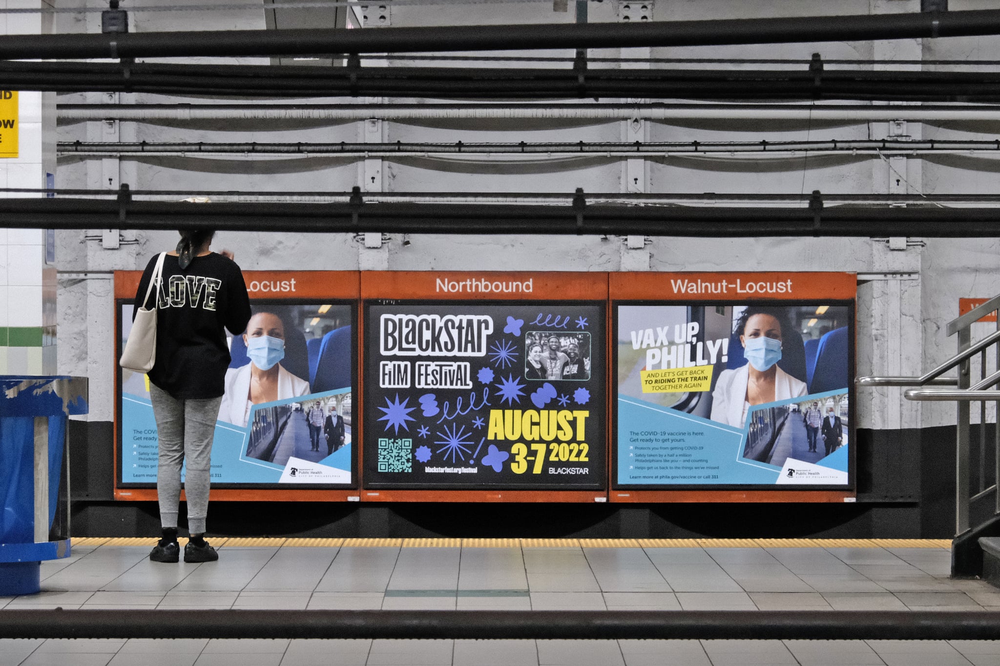 A photo of a Philly subway station. A woman is standing in front a three sheet poster display. The poster in the middle is advertising BlackStar Film Festival.