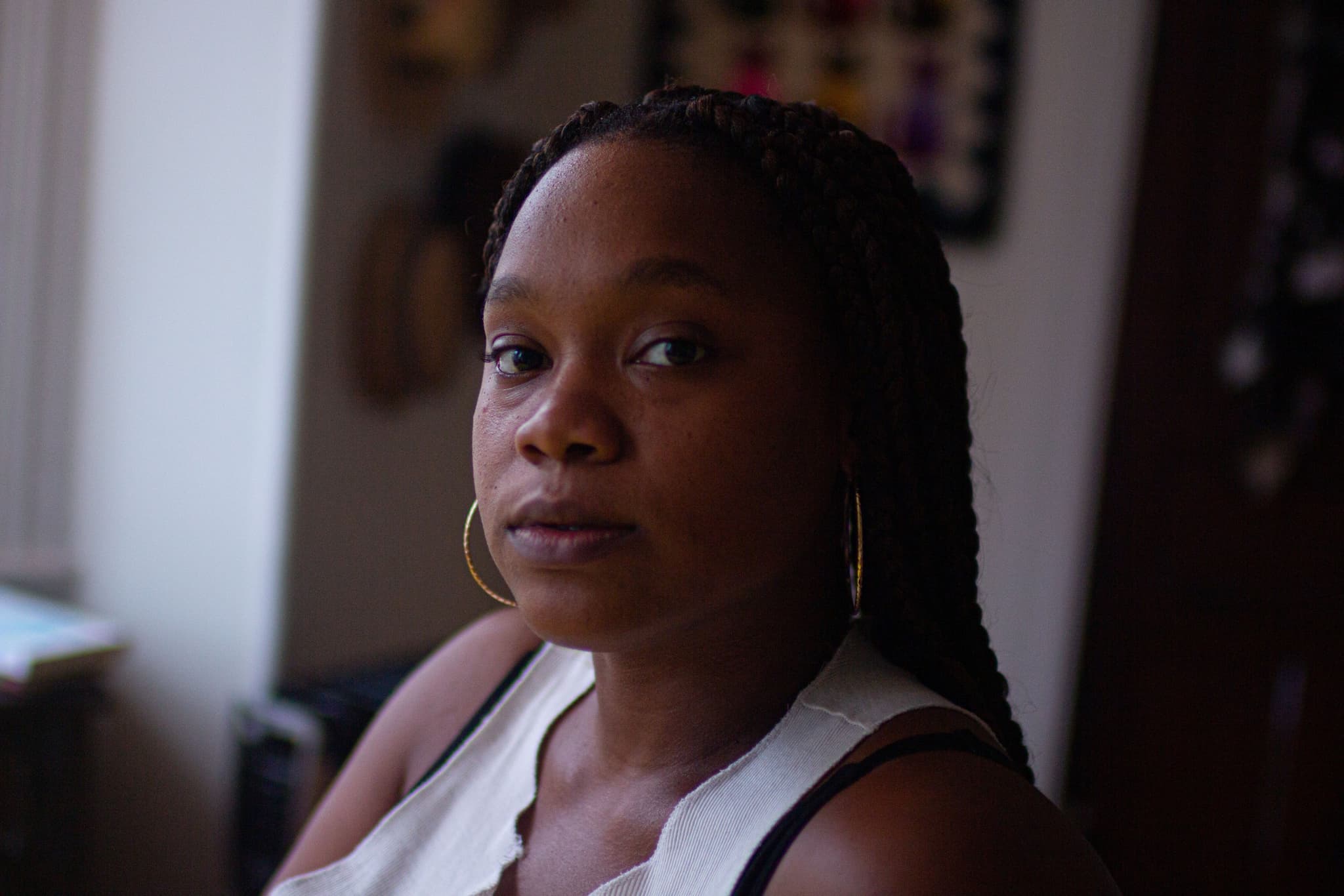 Headshot of Tshay Williams shows her sitting in a living room space. Various pieces of art can be seen in the background though they are blurry. The focus is on Williams, who is turning her head to look directly at the camera. She wears a white tank top with a deep V neckline and golden hoop earrings. Her hair is in long braids that fall down her back.