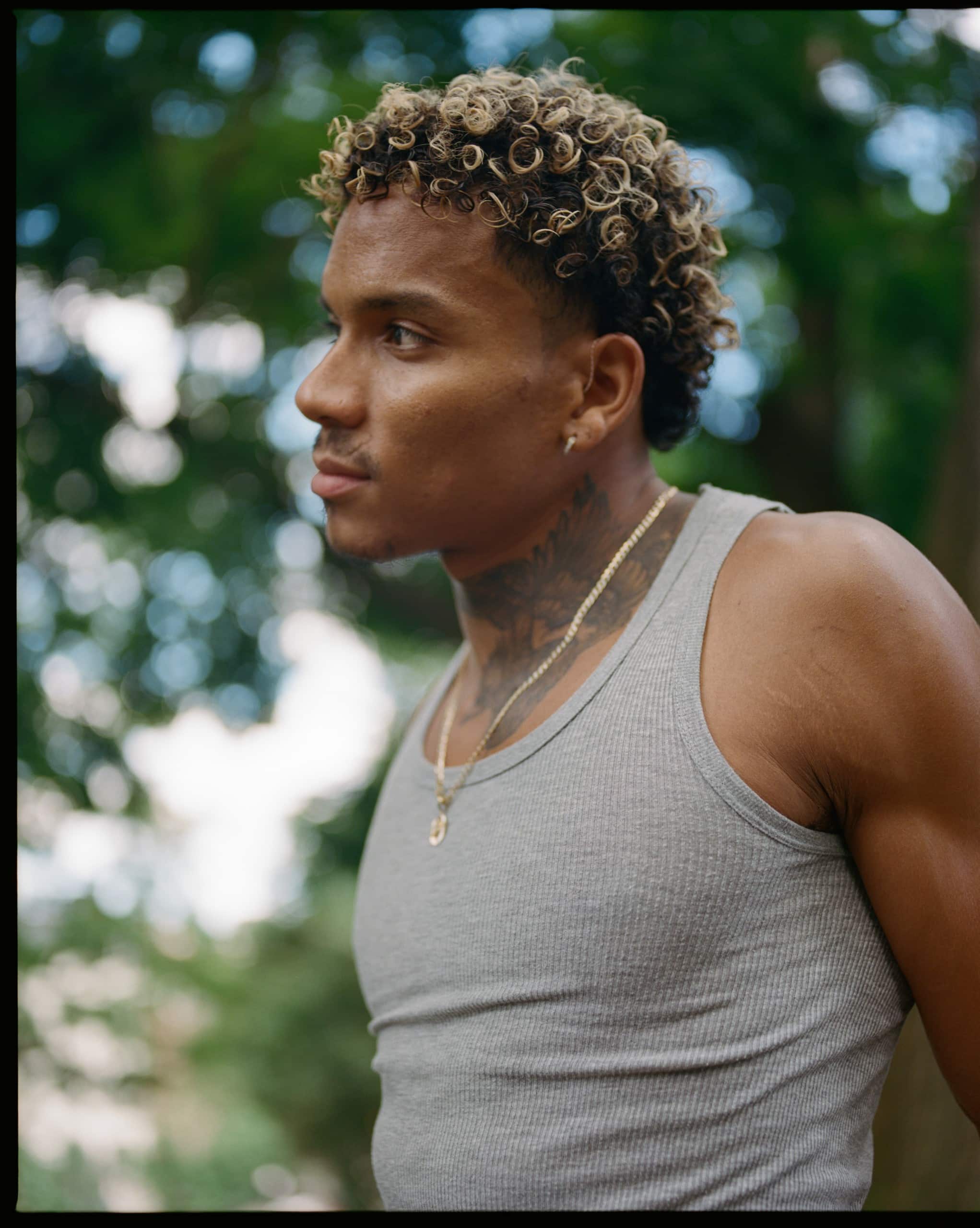 A photo of a Black person in side profile from the mid-waist up, looking to the left while wearing a gray tank top and a gold chain. Their arms are placed behind, as if they might be held behind their back. In the distance we can make out the blurry outline of some green trees.