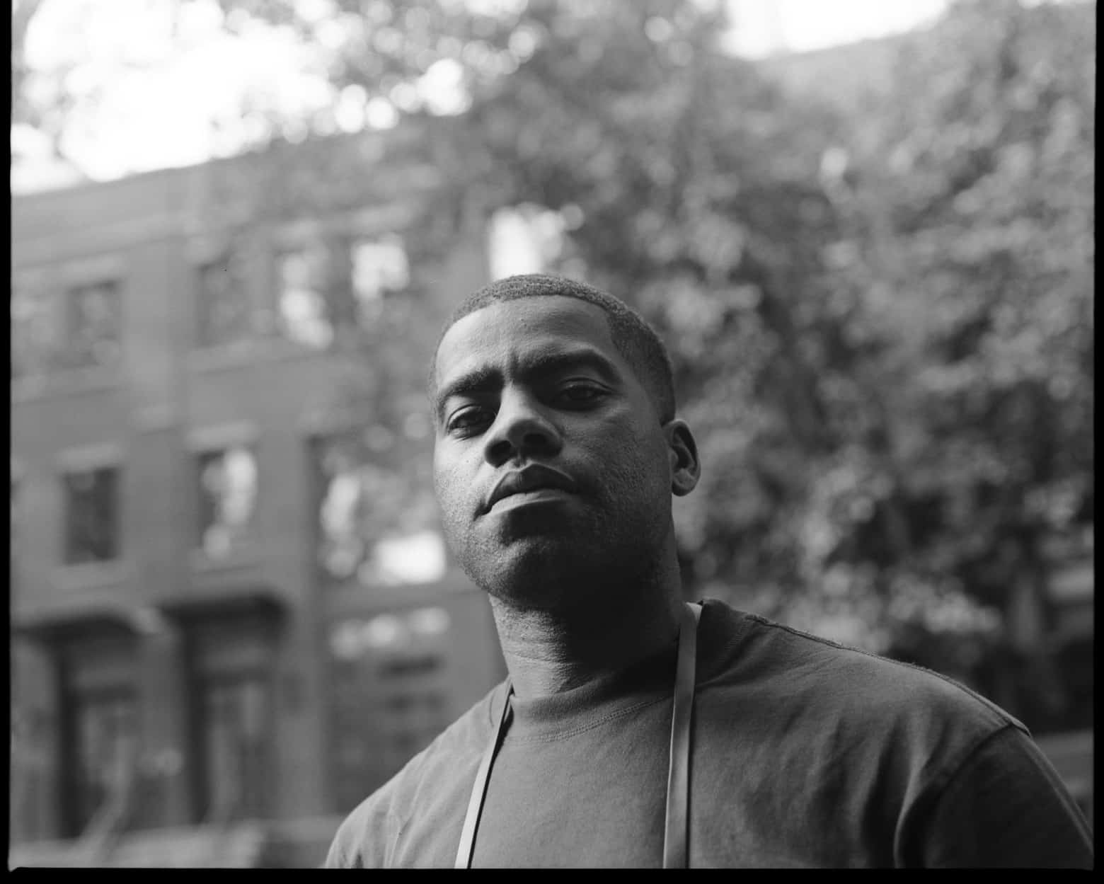 A Black person looks seriously at the camera, standing in front of a brick building and a tree which are both out of focus.