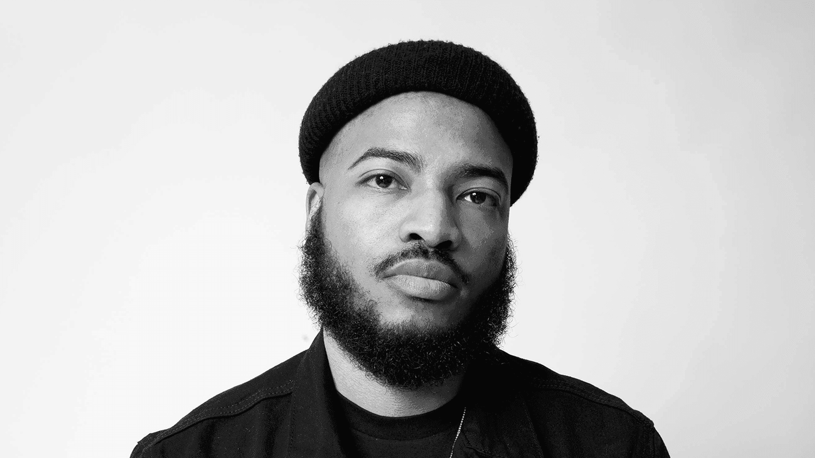 A photo of Darol Olu Kae in black and white, looking directly at the camera, wearing a black beanie hat and a black shirt.
