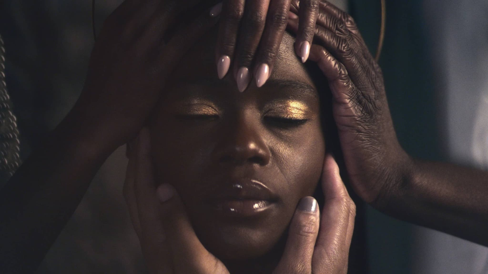 A Black face is centered, with their eyes closed, while a number of other Black hands are touching, caressing, holding their face.