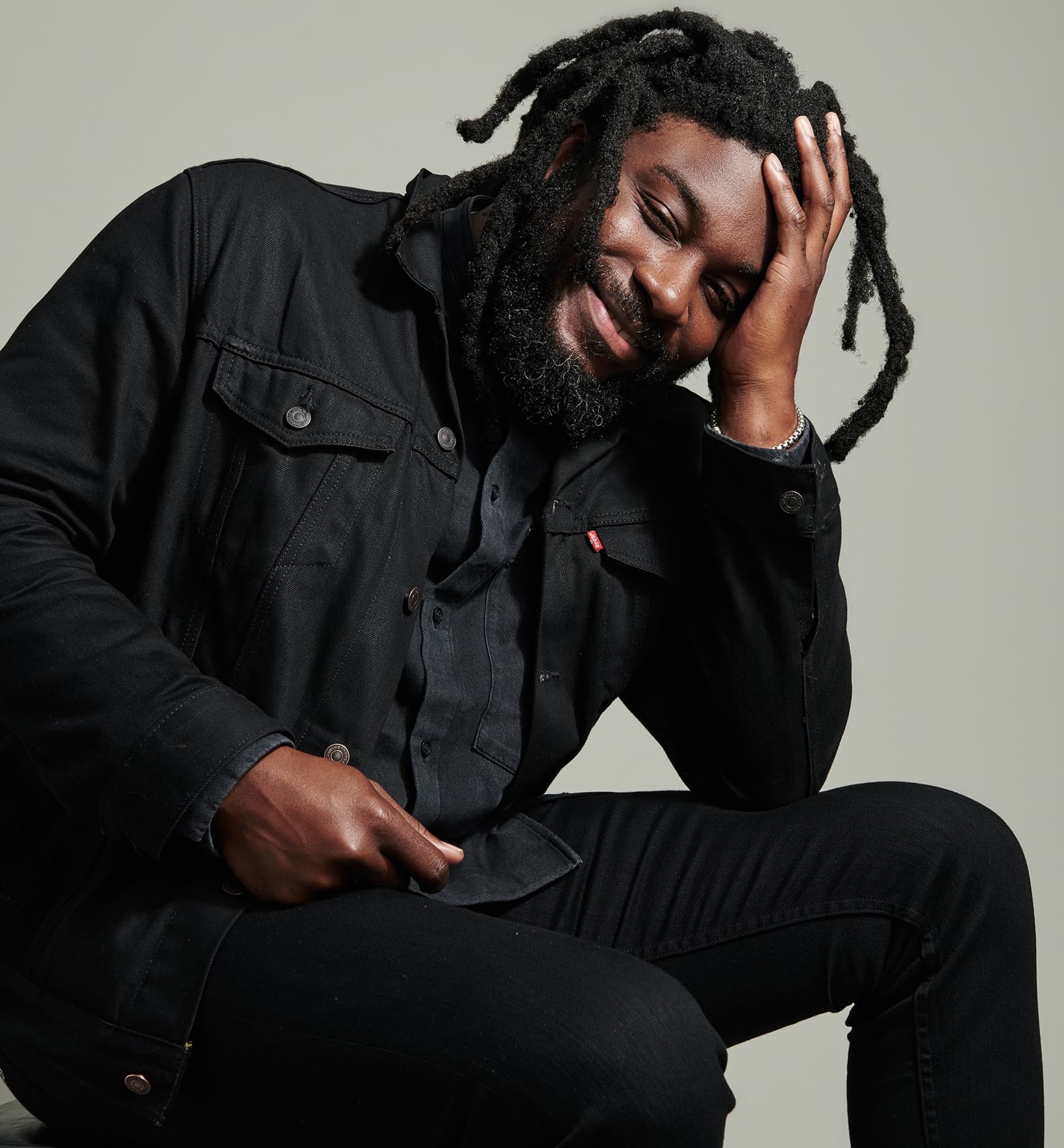 A photo of Jason Reynolds shows him wearing all black and smiling brightly towards the camera while resting his head in his left hand.