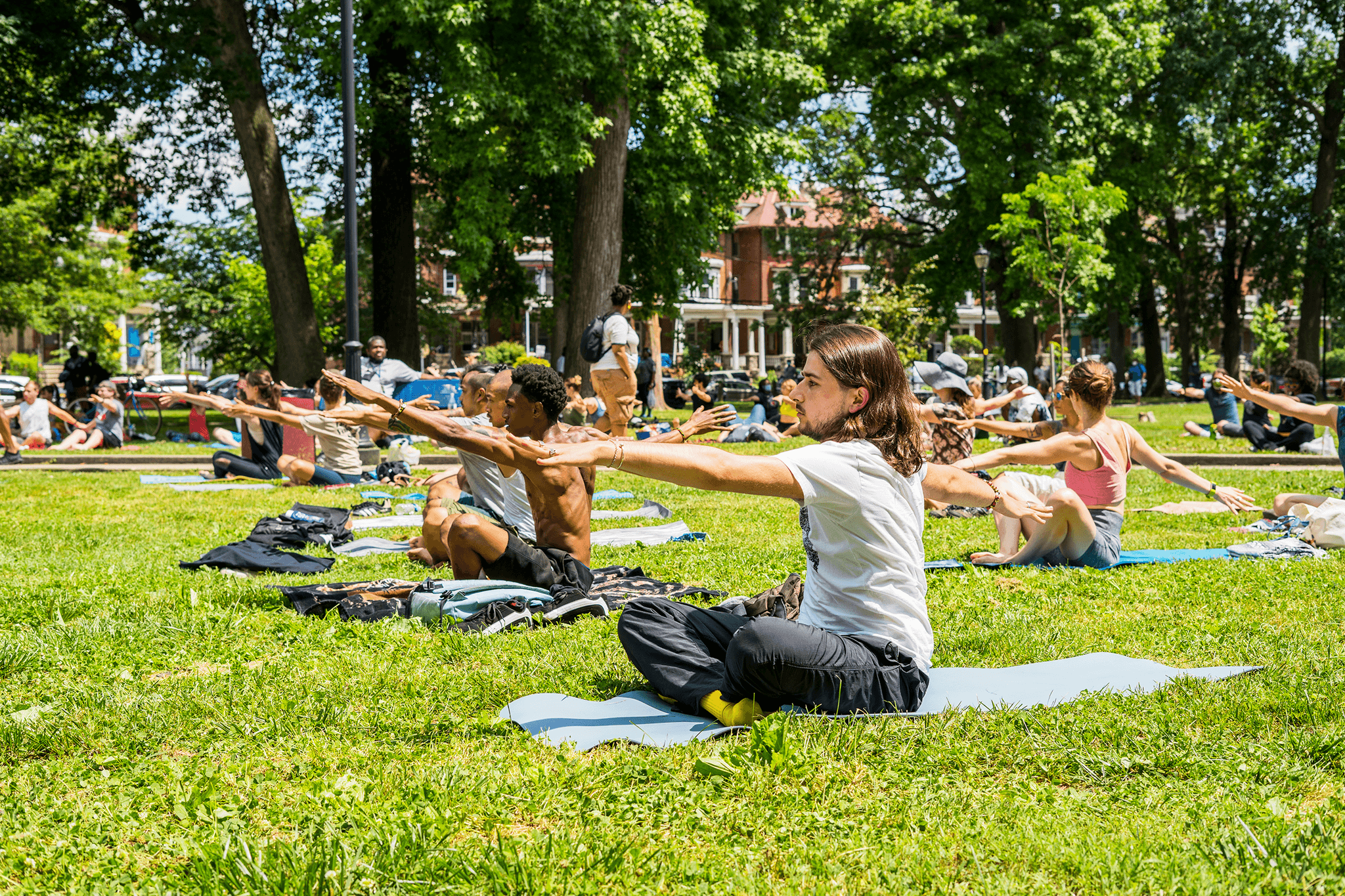 Outdoor photo of a group of people sitting on a large grassy area. They sit on yoga mats and stretch their hands out in front of them.