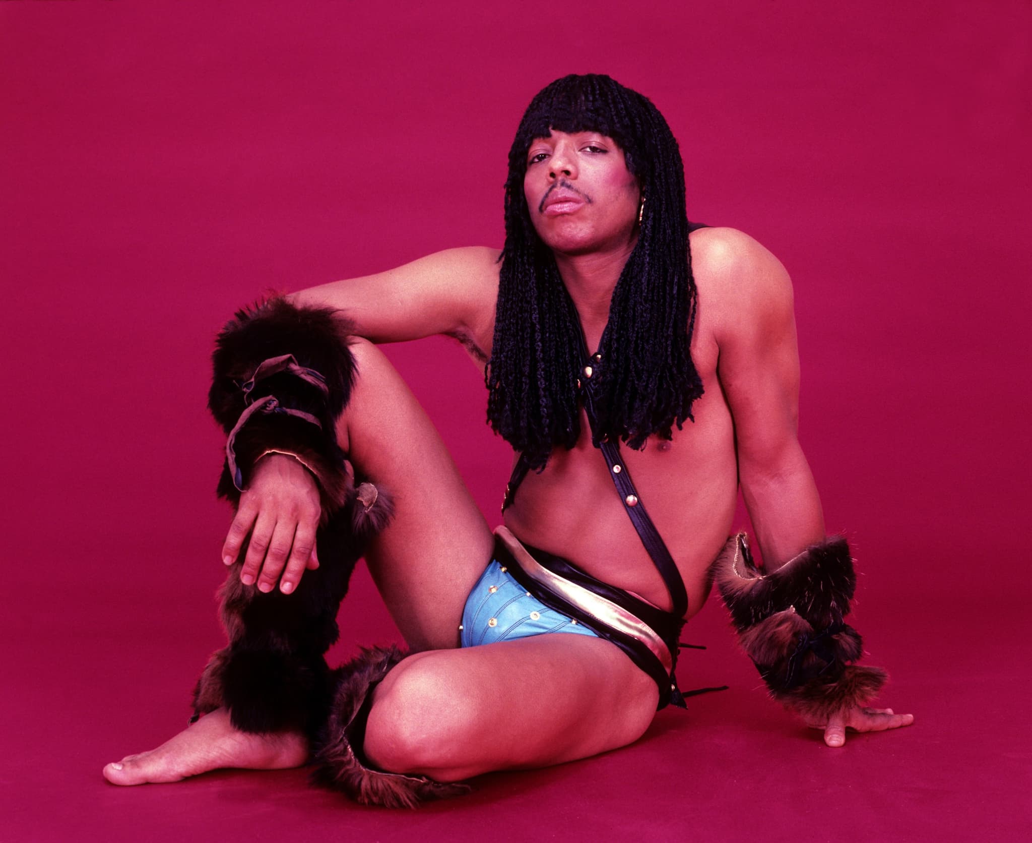 A photo of Rick James shows him posing while sitting on the floor in only underwear, his right arm drapped over his right knee, which is raised off the ground. The background is all red.