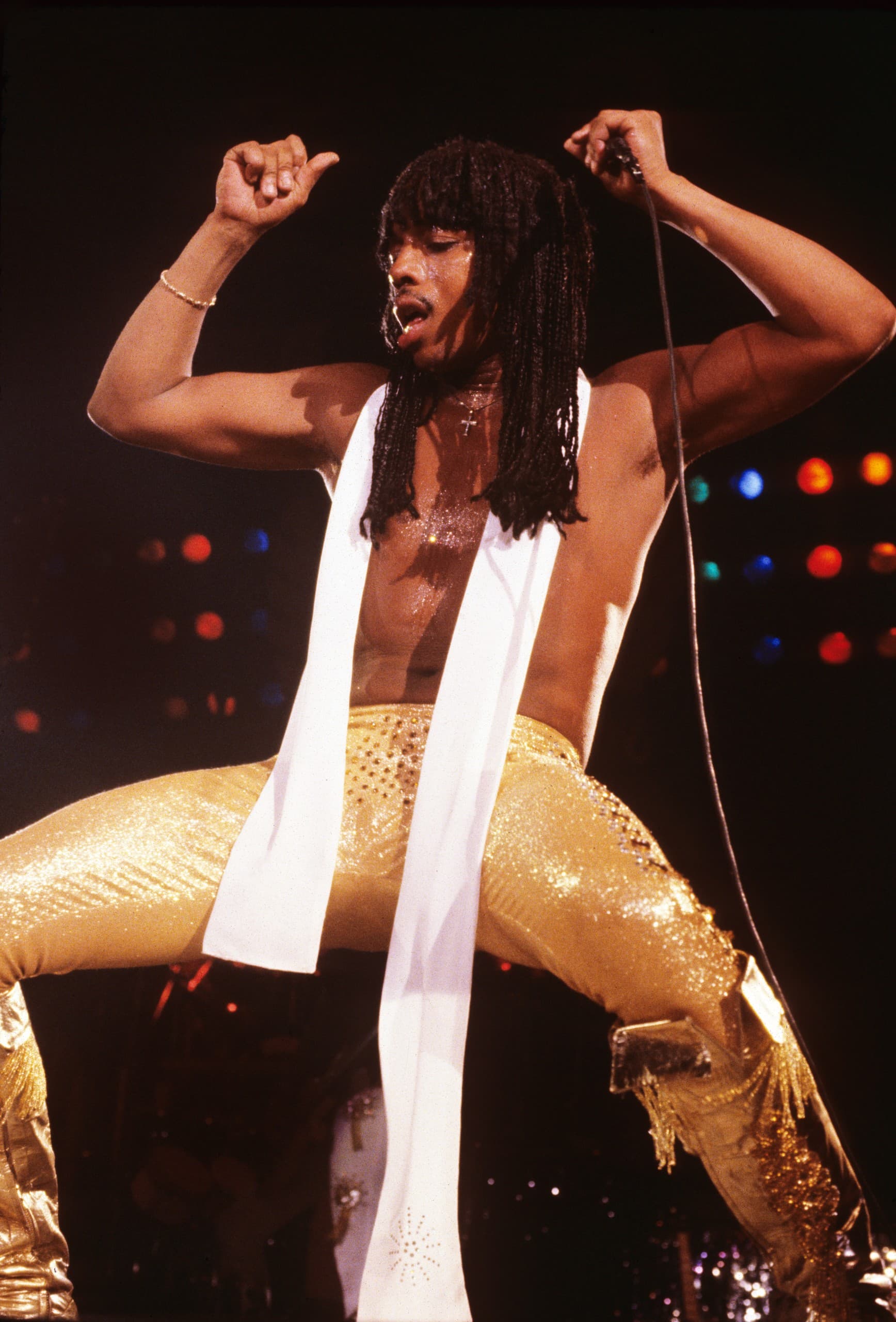 Rick James performs on stage in shiny yellow leather pants and a towel around his neck, shirtless, his arms raised in mid dance.