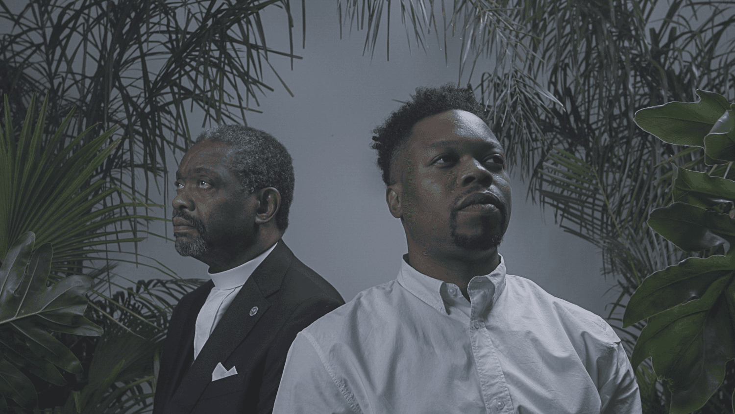 Two Black people look in opposite directions while standing against a gray background, some foliage is visible at the edges.