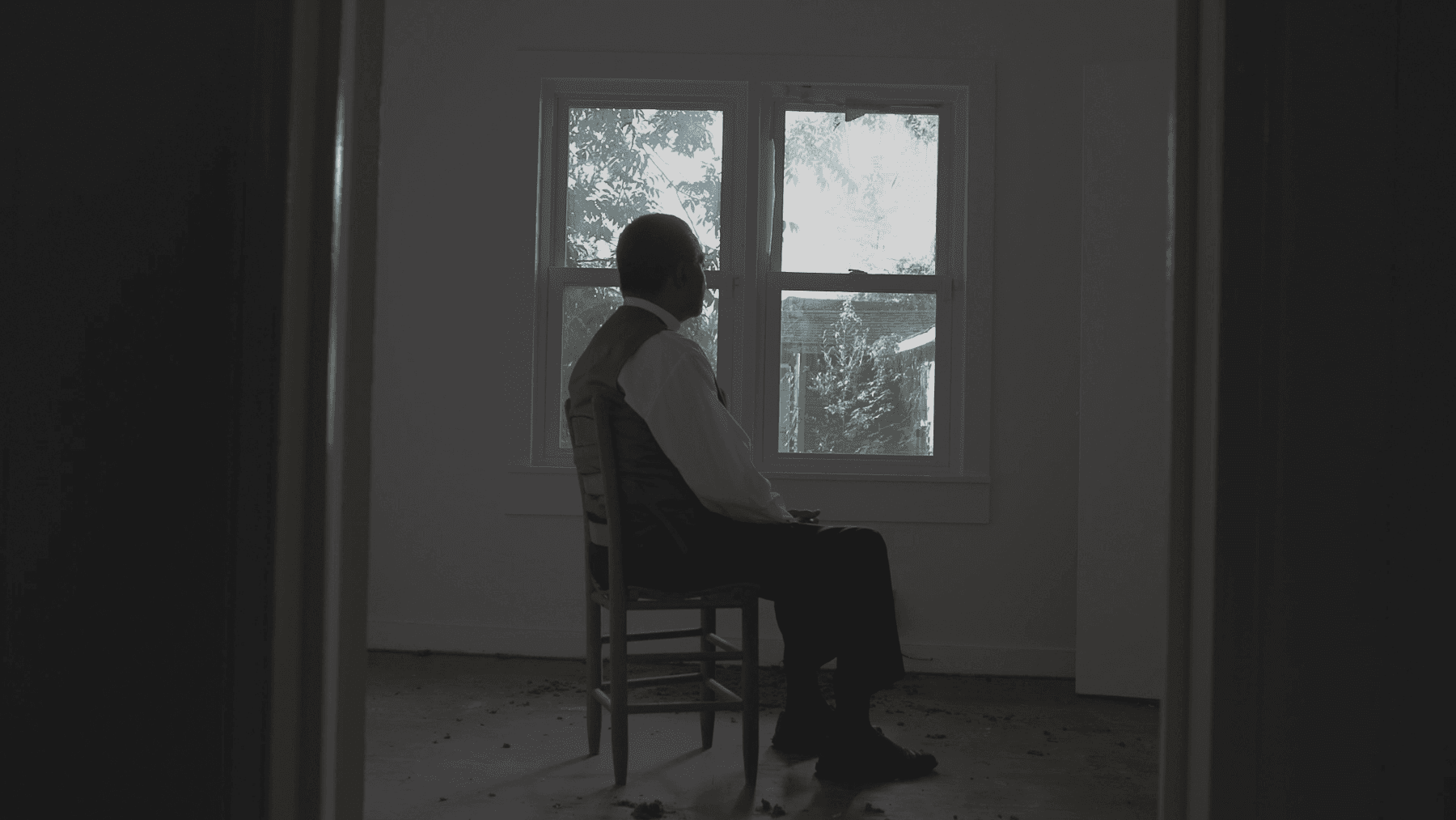 A Black person sits inside a house in shadow, but behind them is an open window which indicates that it is day outside.