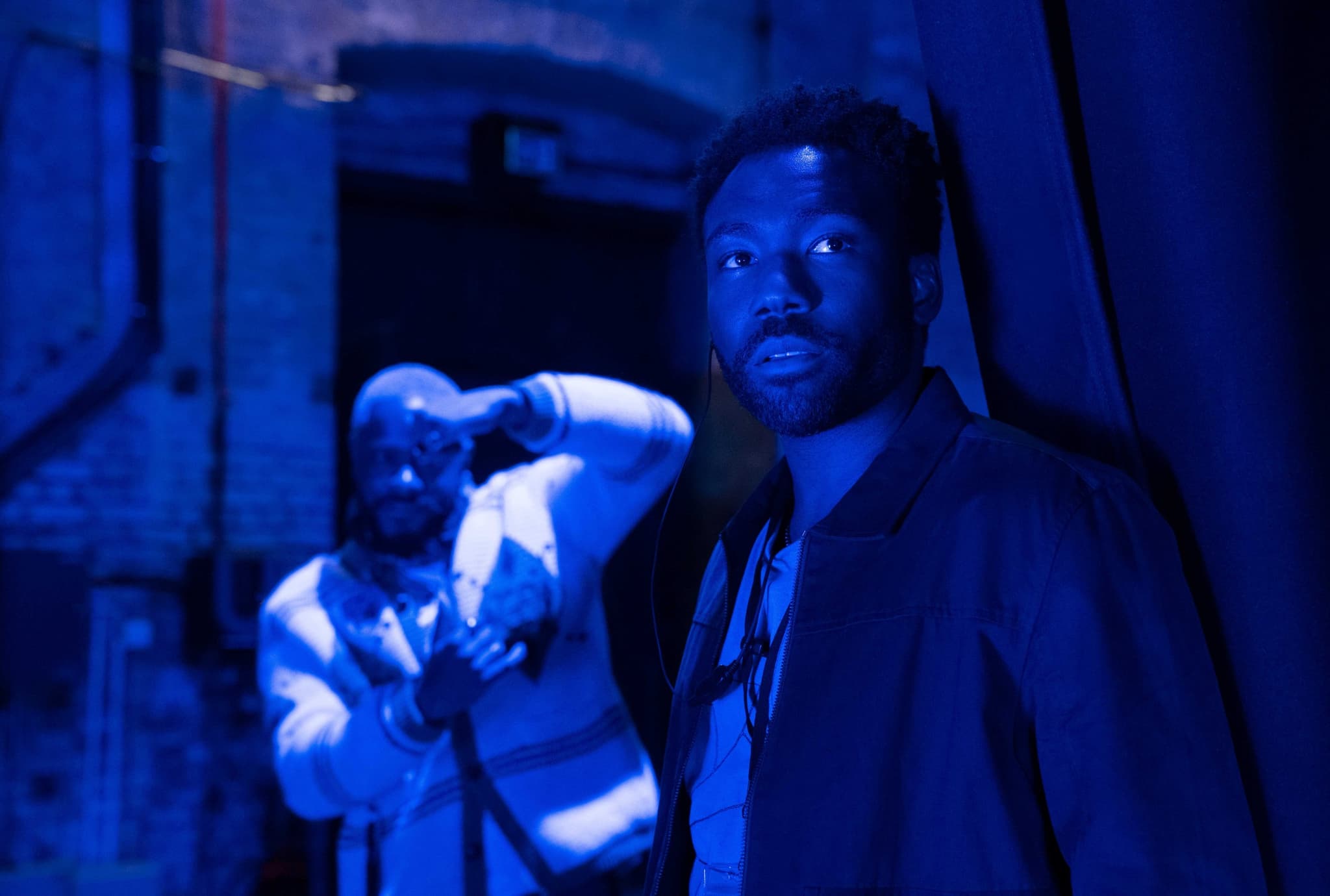 Two Black men stand facing the viewer in a dark, blue-lit space. Donald Glover stands in the foreground, looking off frame, while LaKeith Stanfield stands in the background, with a hand framing his left eye.