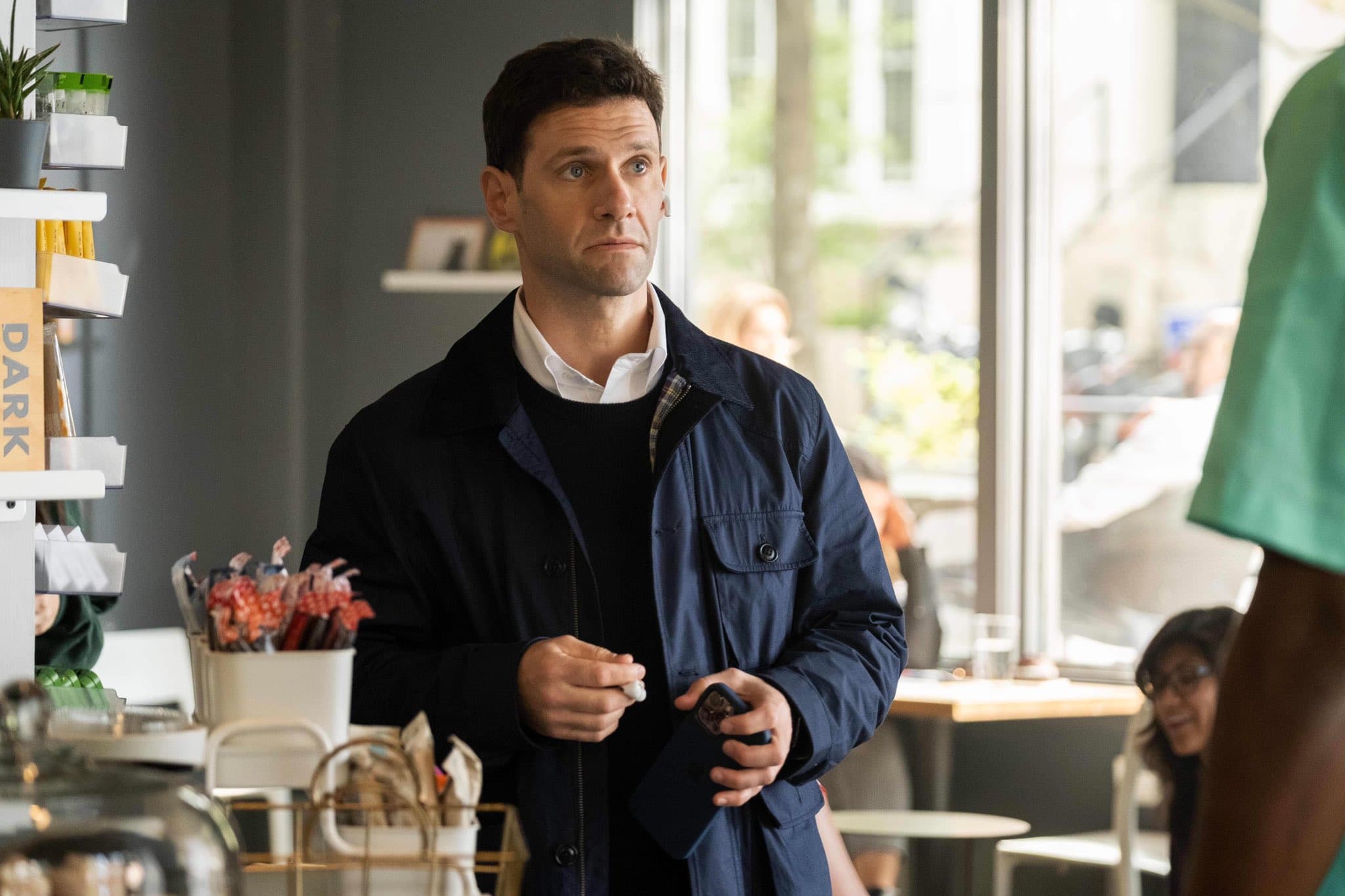 A bewildered white man stands in a coffee shop, wearing a navy jacket and clutching an iPhone.