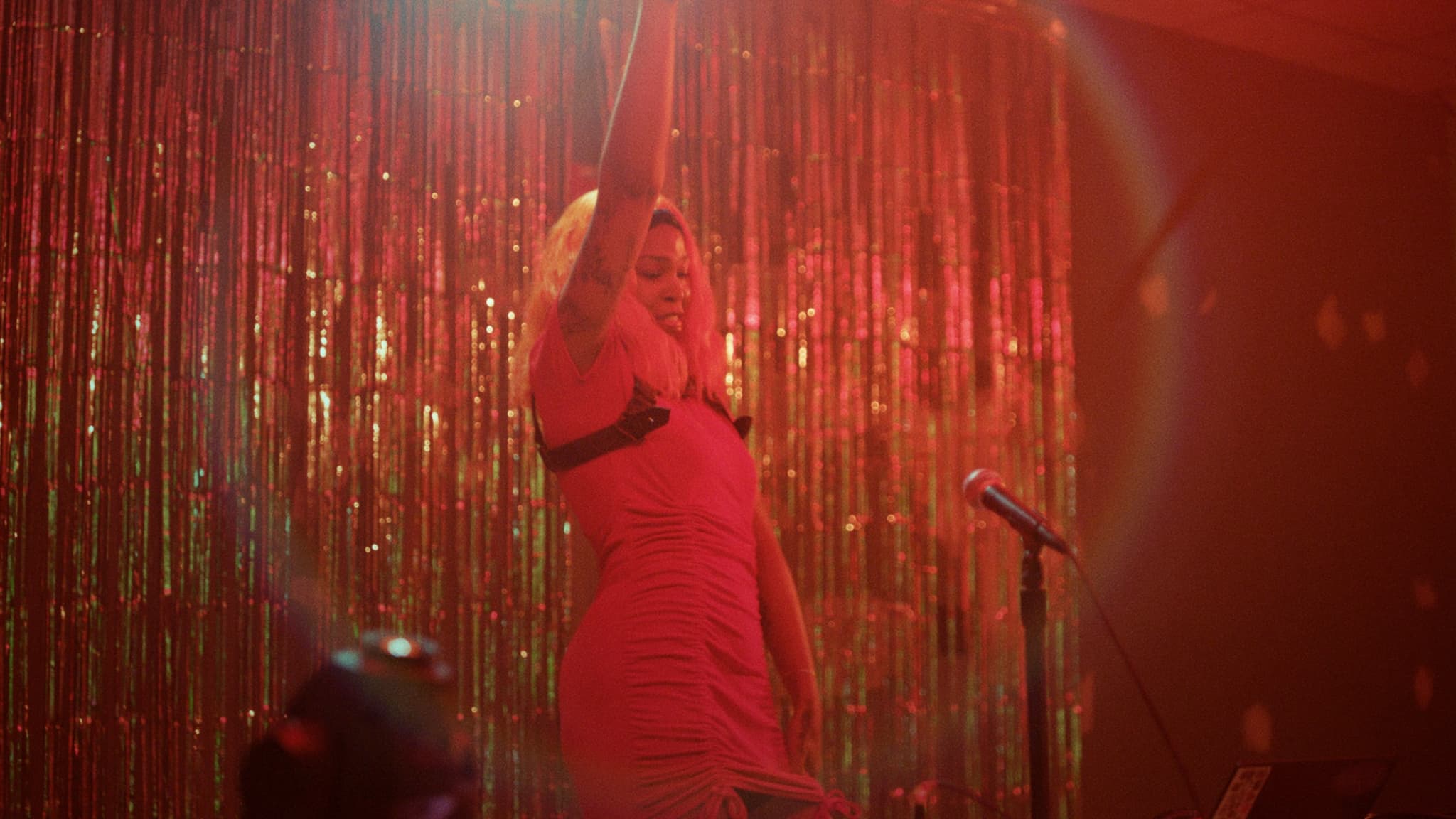 A Black person dances on stage - a microphone in front of them -against a red hued background that shimmers