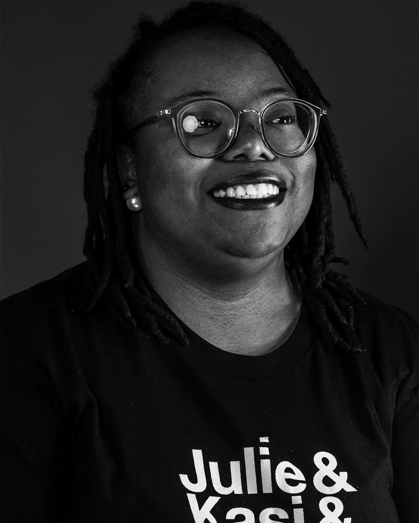 A black and white headshot of Tamira Moore shows her smiling and looking off screen. She wears dark lipstick and large hoop earrings as well as eyeglasses.