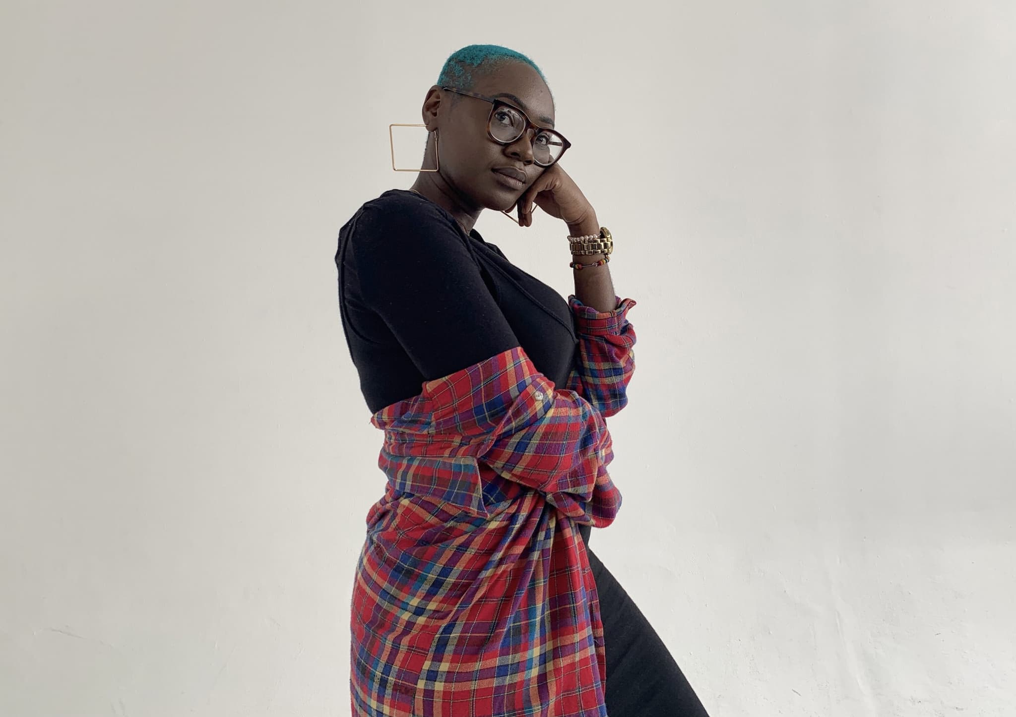 A photo of Animah Danquah shows her posing in front of a white background. She wears a red flannel shirt that is slightly falling off her person. Her hair is cropped short to her head and dyed bright blue. She lifts a hand and presses it against her cheek while looking into the camera.