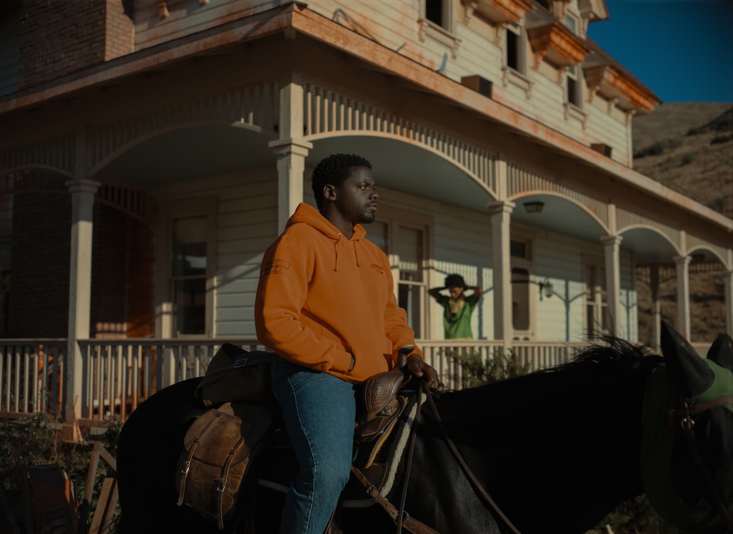 A still from Nope shows Daniel Kaluya's character sitting on a horse, looking off to the right, wearing a bright orange sweatshirt. In the background, on the porch of an old house, is Keke Palmer's character looking exasperated.