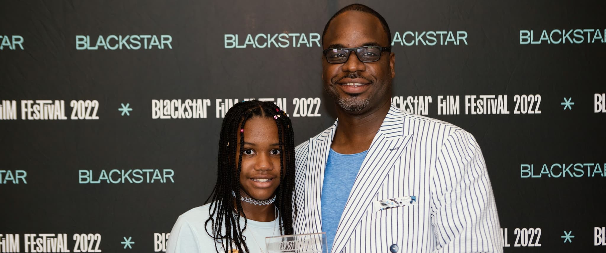 A photo of Bria and Bilal Motley, a Black father-daughter duo, they're standing in-front of a step-and-repeat that says "BlackStar Film Festival 2022." They are holding an award.